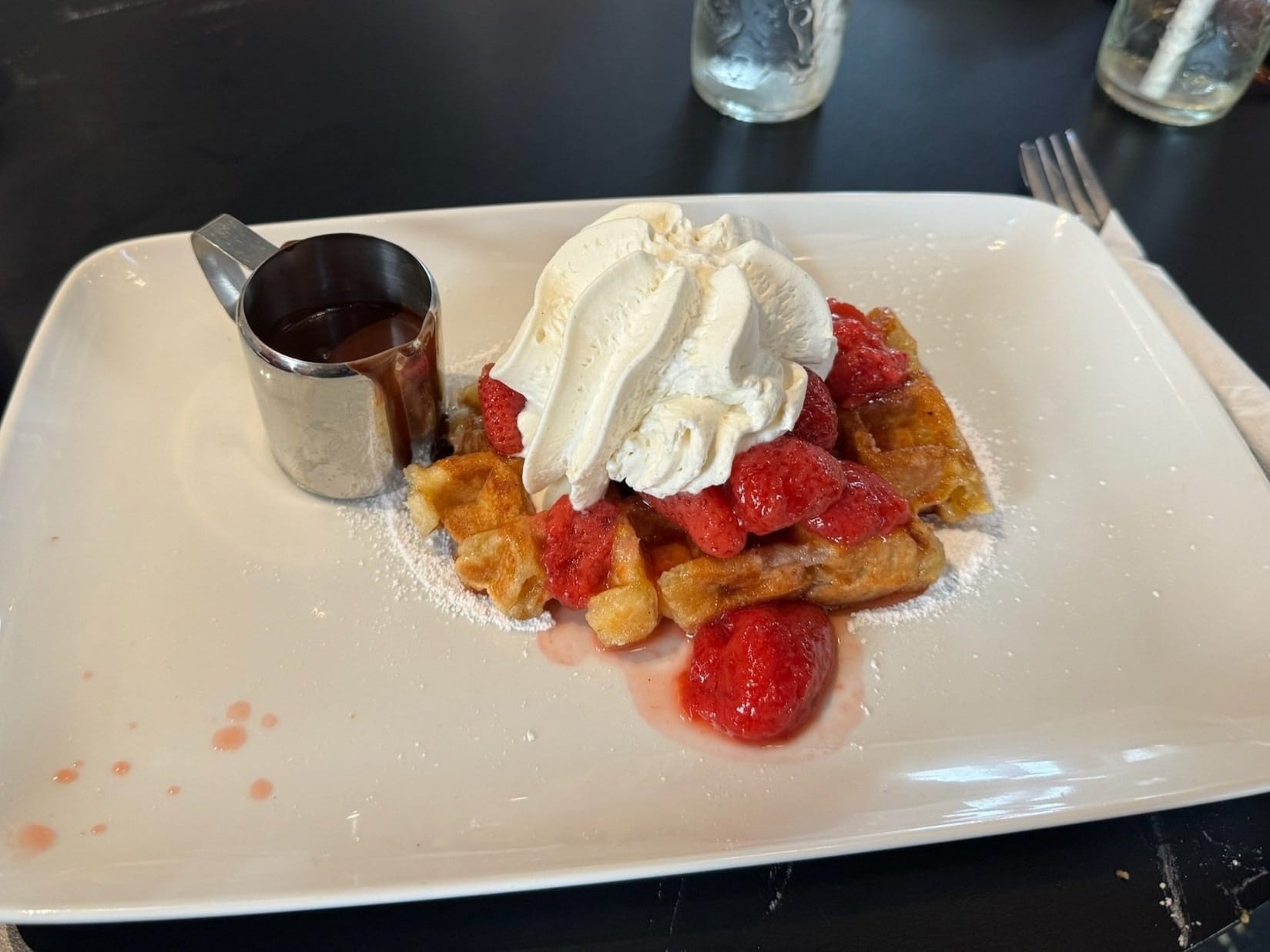 A delicious plate of waffles topped with strawberries, whipped cream, and a side of syrup.