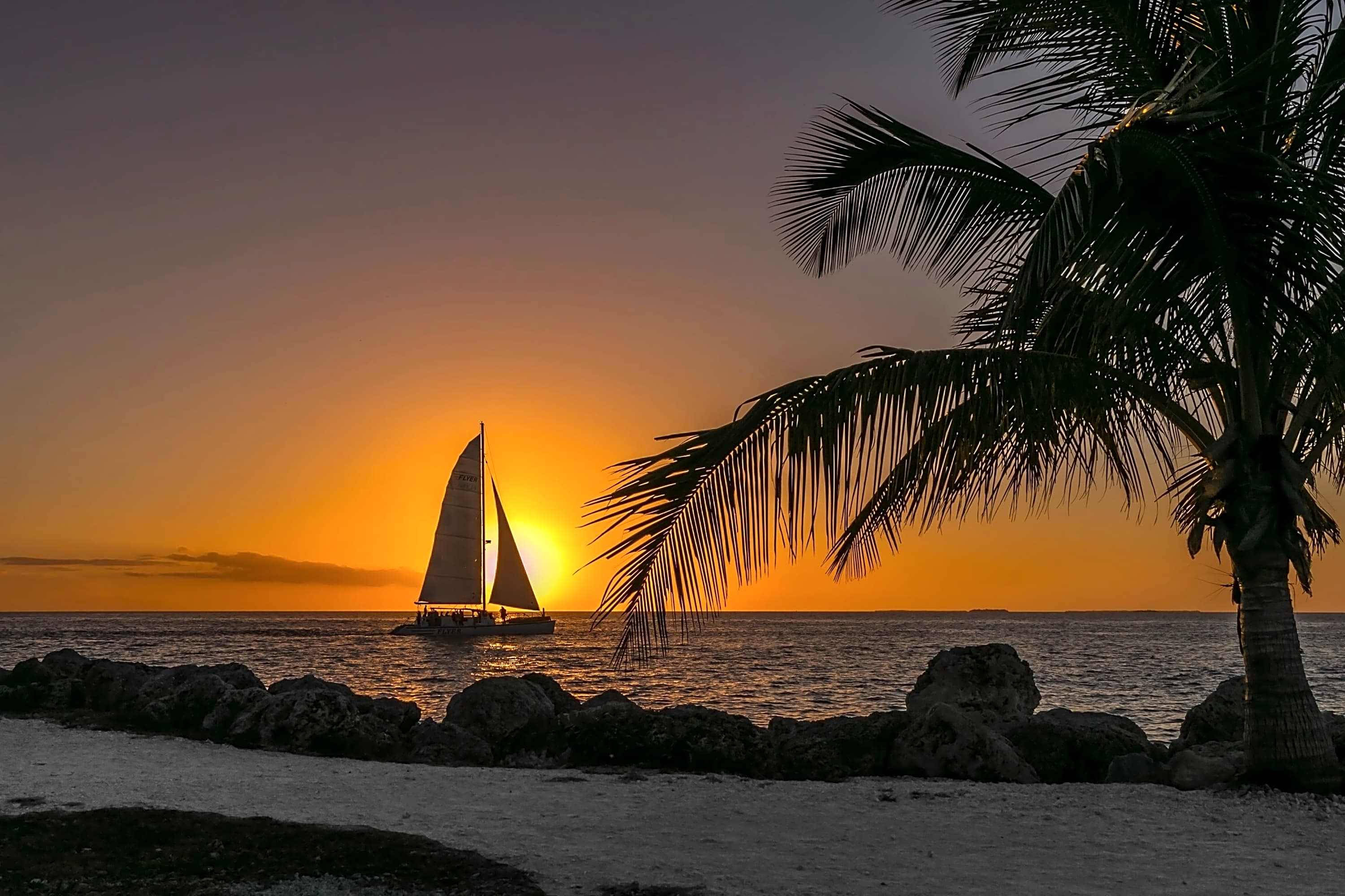 A sailboat sails along the Key West shoreline with the sun setting behind it