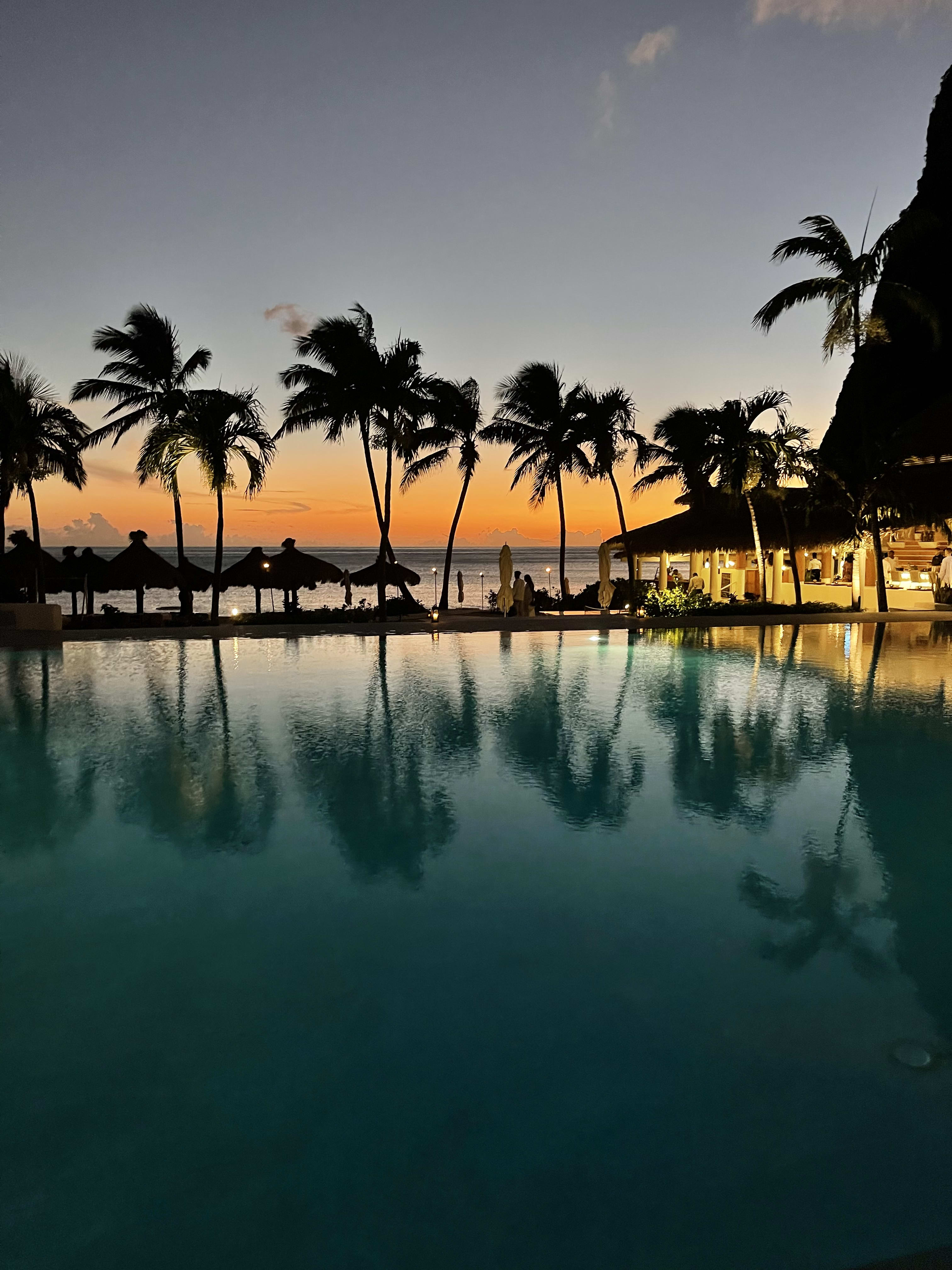 Beautiful view of a pool surrounded by palm trees and cabanas on the beach at sunset