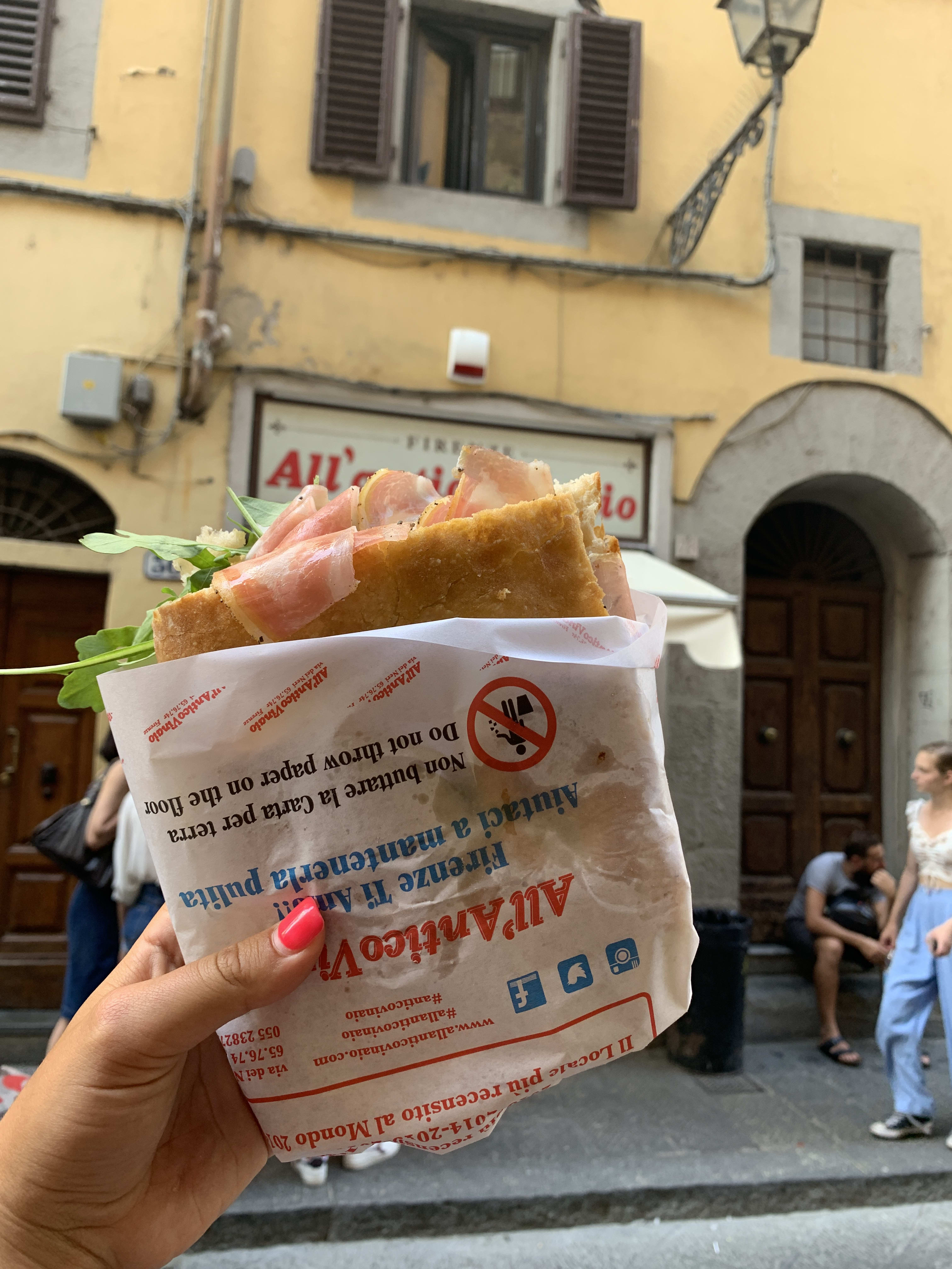 Photo of a someone holding up a focaccia sandwich in front of an old, yellow house