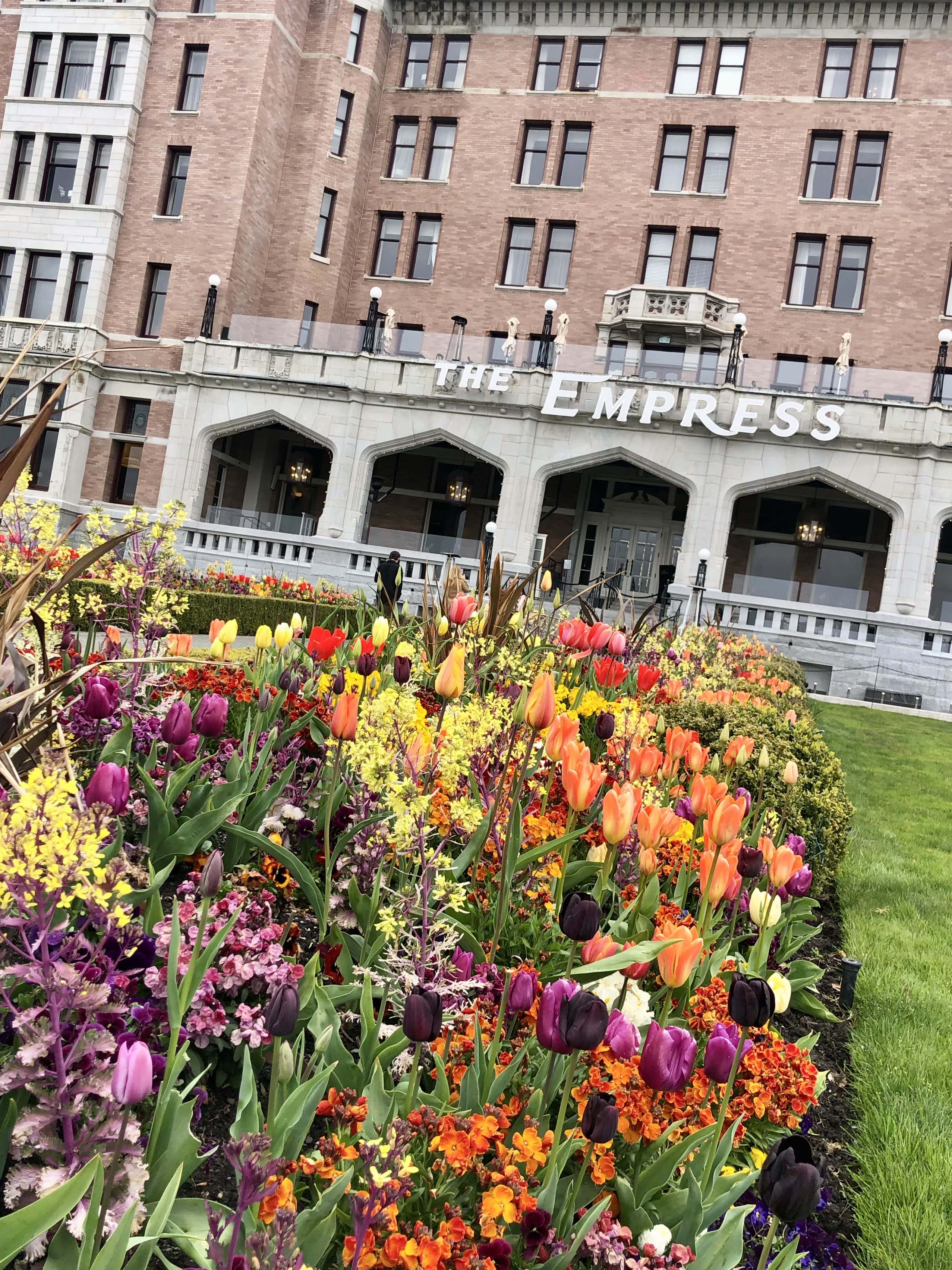 A bed of colorful flowers on a grassy square in front of a red and white building with The Empress sign.