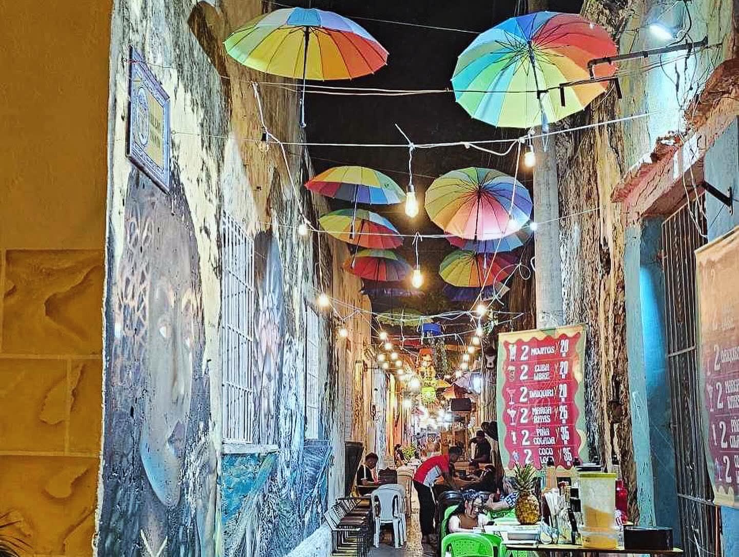 A lively street, lit up at night and decorated with murals and colorful umbrellas hanging from above.