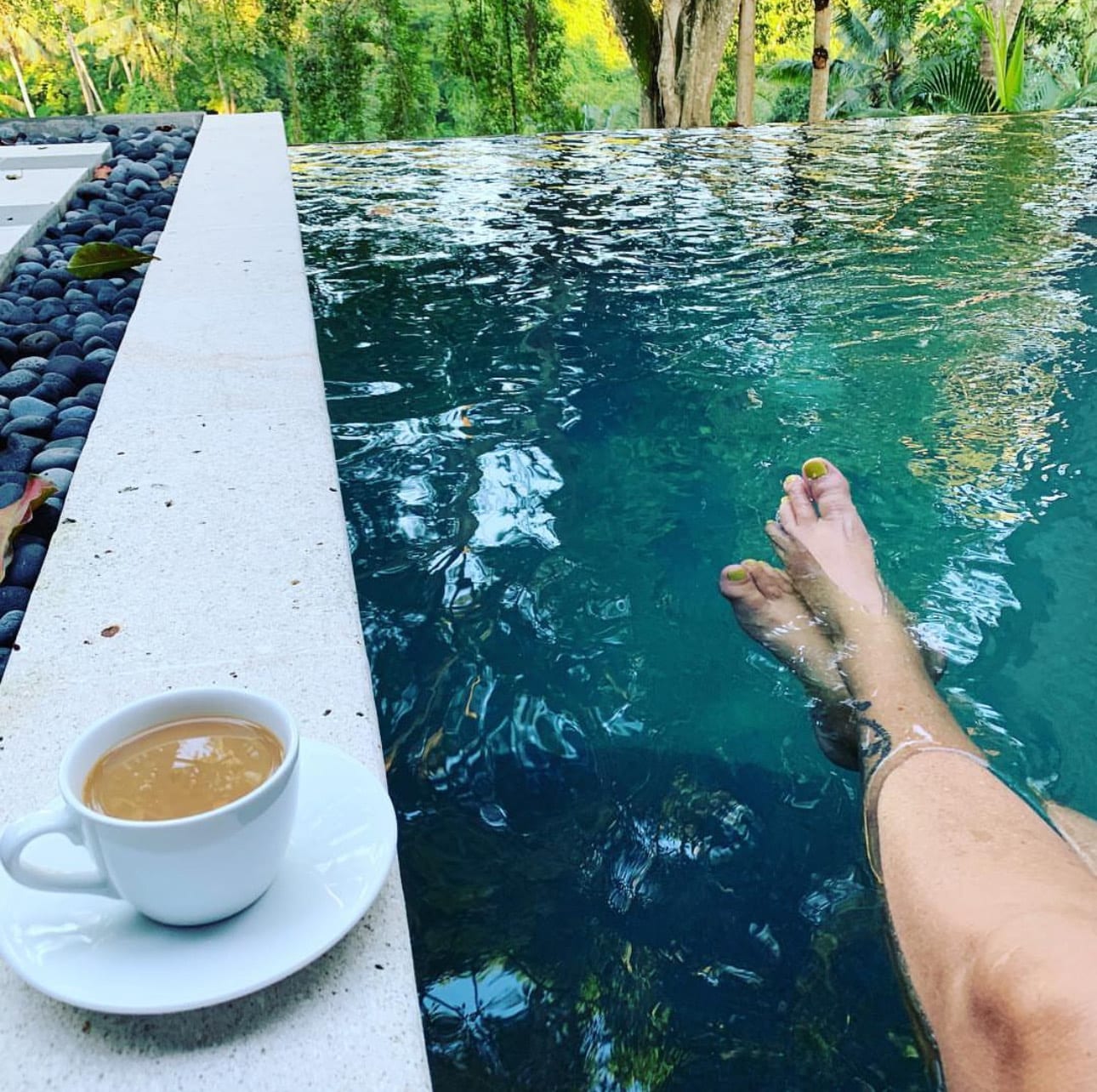 Laura's legs in an infinity pool with a cup of coffee on the ledge overlooking trees.