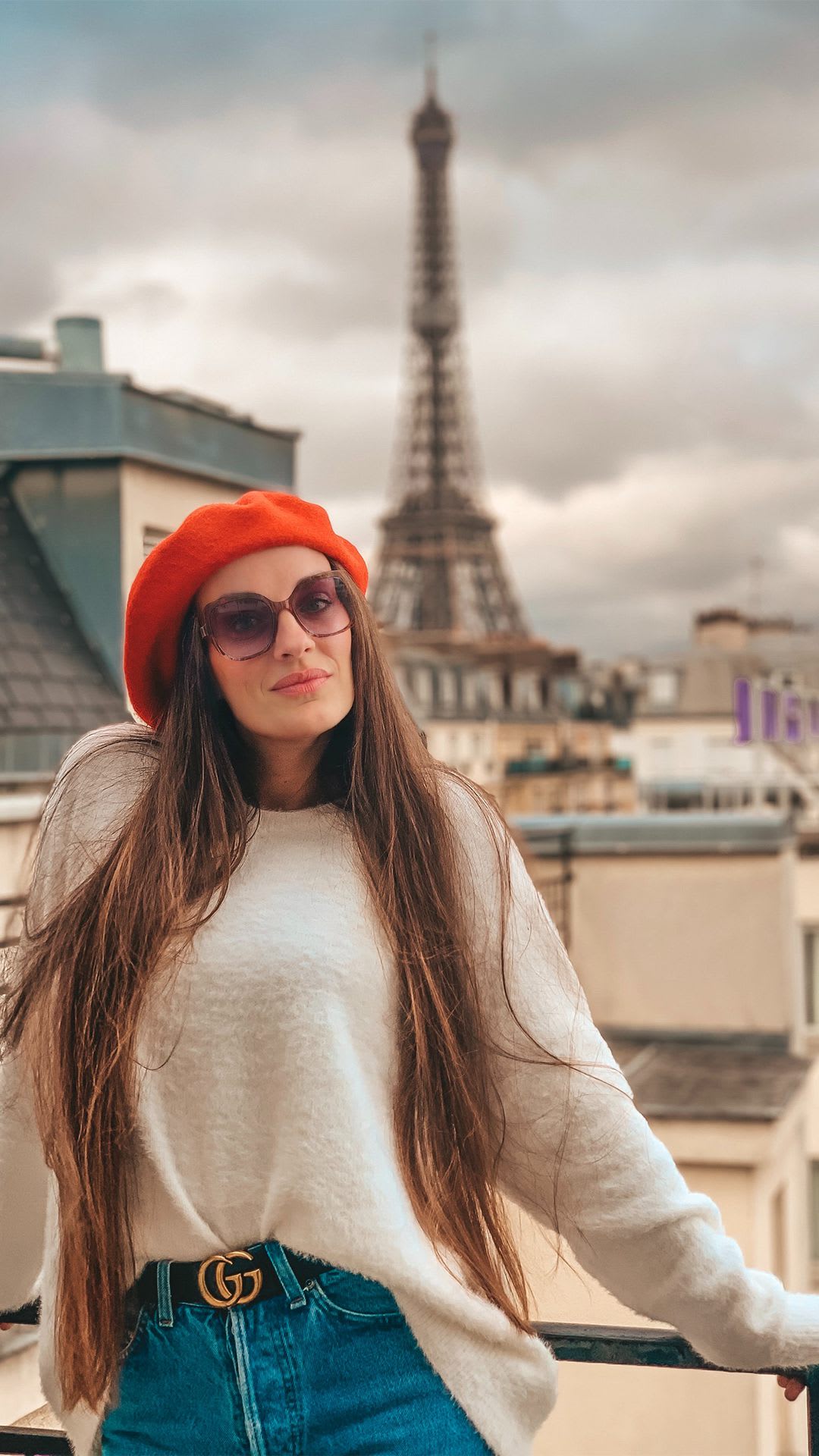 Britnee in a white sweater and red beret posing for a photo on a balcony with a view of the Eiffel Tower