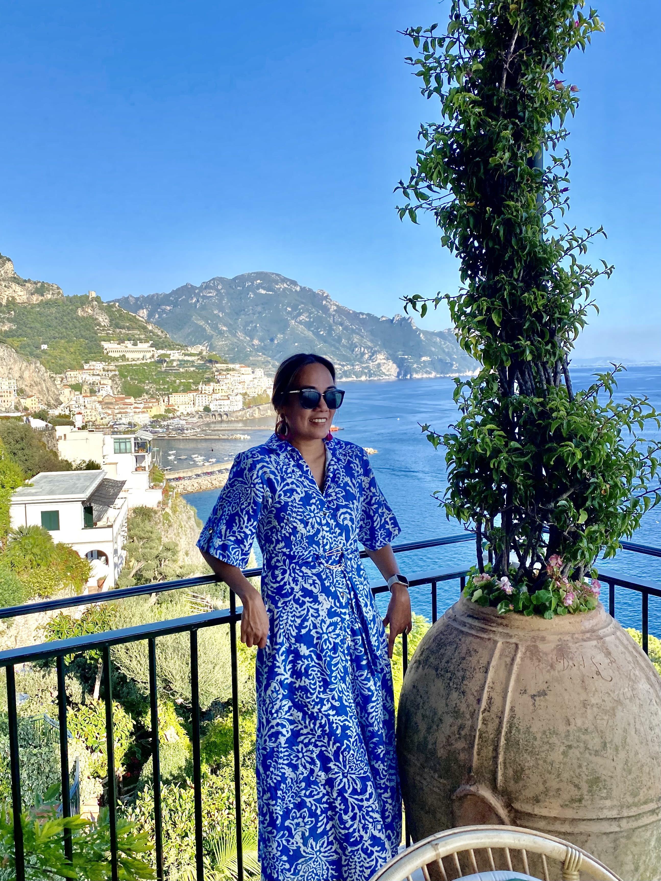 Pam standing on a balcony in a blue dress with beautiful view of the sea and the Amalfi Coast in the background