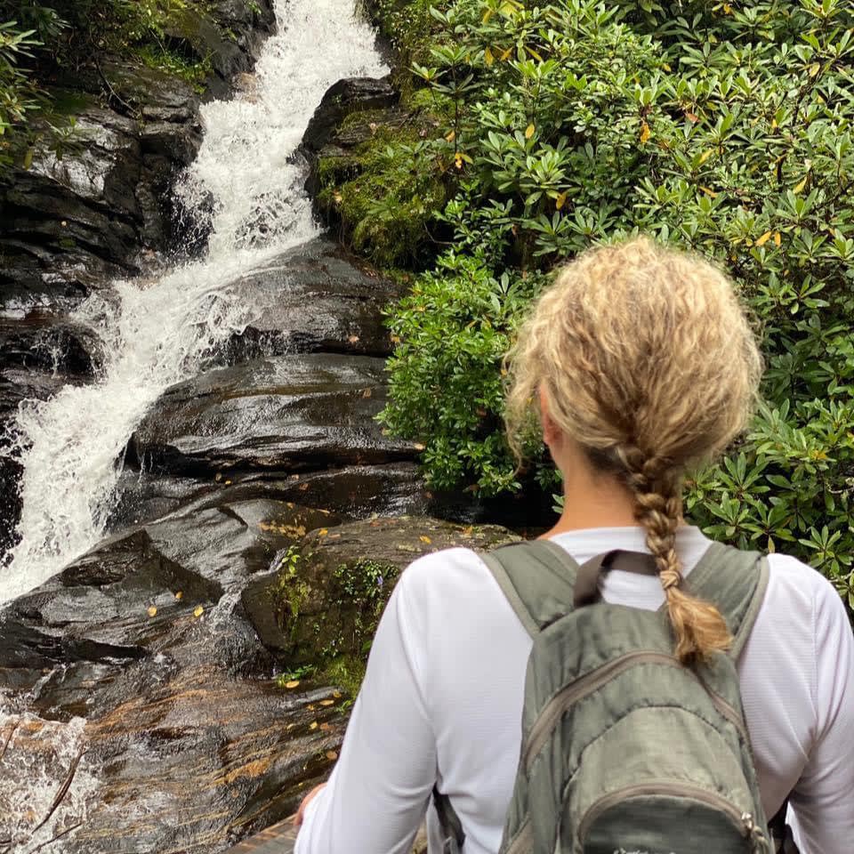 Picture of Melody wearing a braid and facing a cascading waterfall with a green backpack