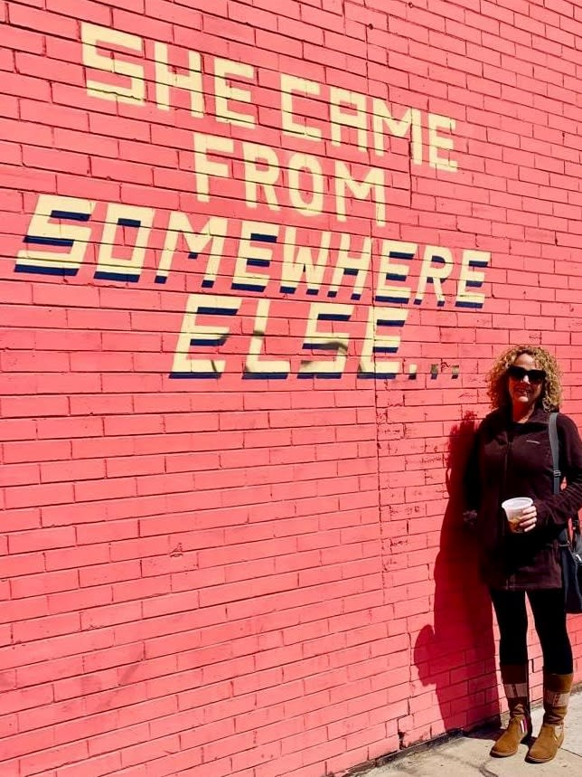Picture of Melody with “she came from somewhere else” written on a pink brick wall