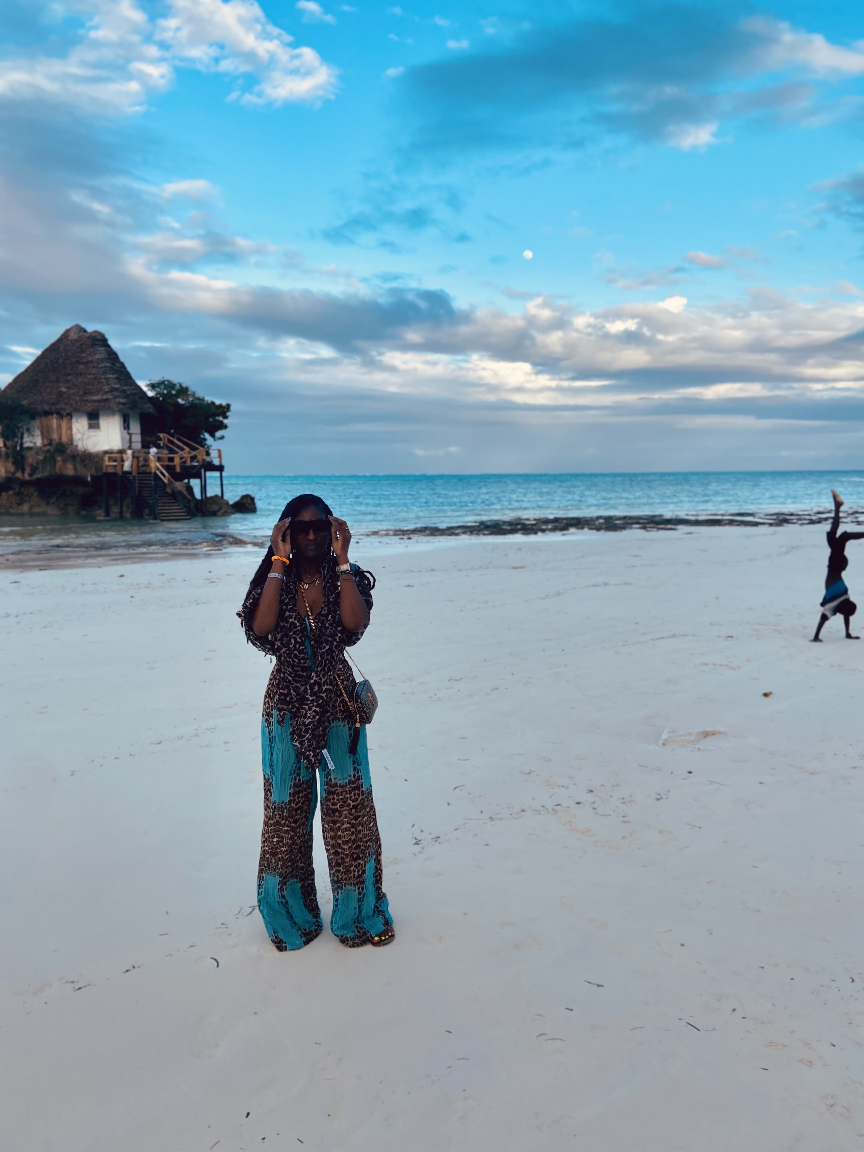 Chardon standing on a white sand beach with an over-the-water bungalow in the background.