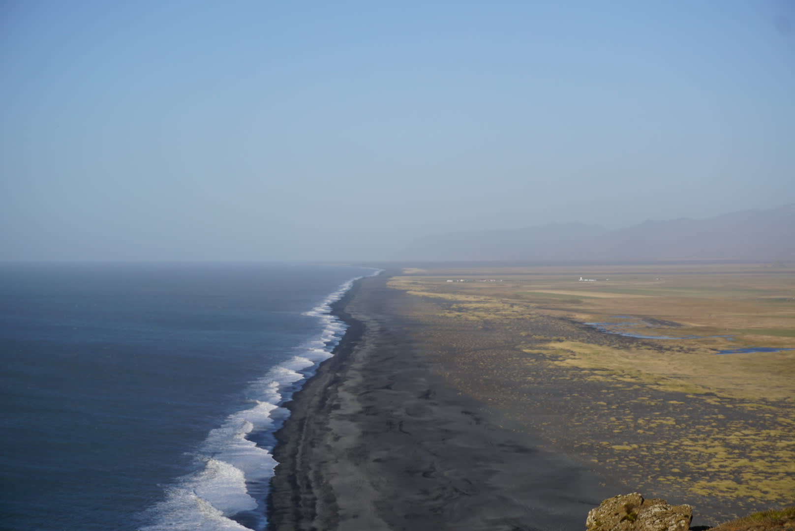 A serene coastal landscape with a dark sandy beach, vast grasslands, and distant mountains under a clear sky.