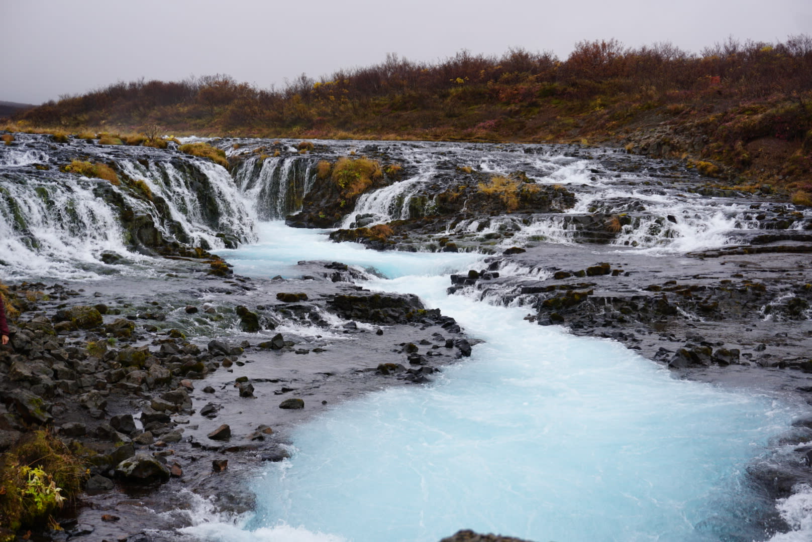 A vibrant waterfall cascades into turquoise waters, surrounded by rocky terrain and autumnal foliage.