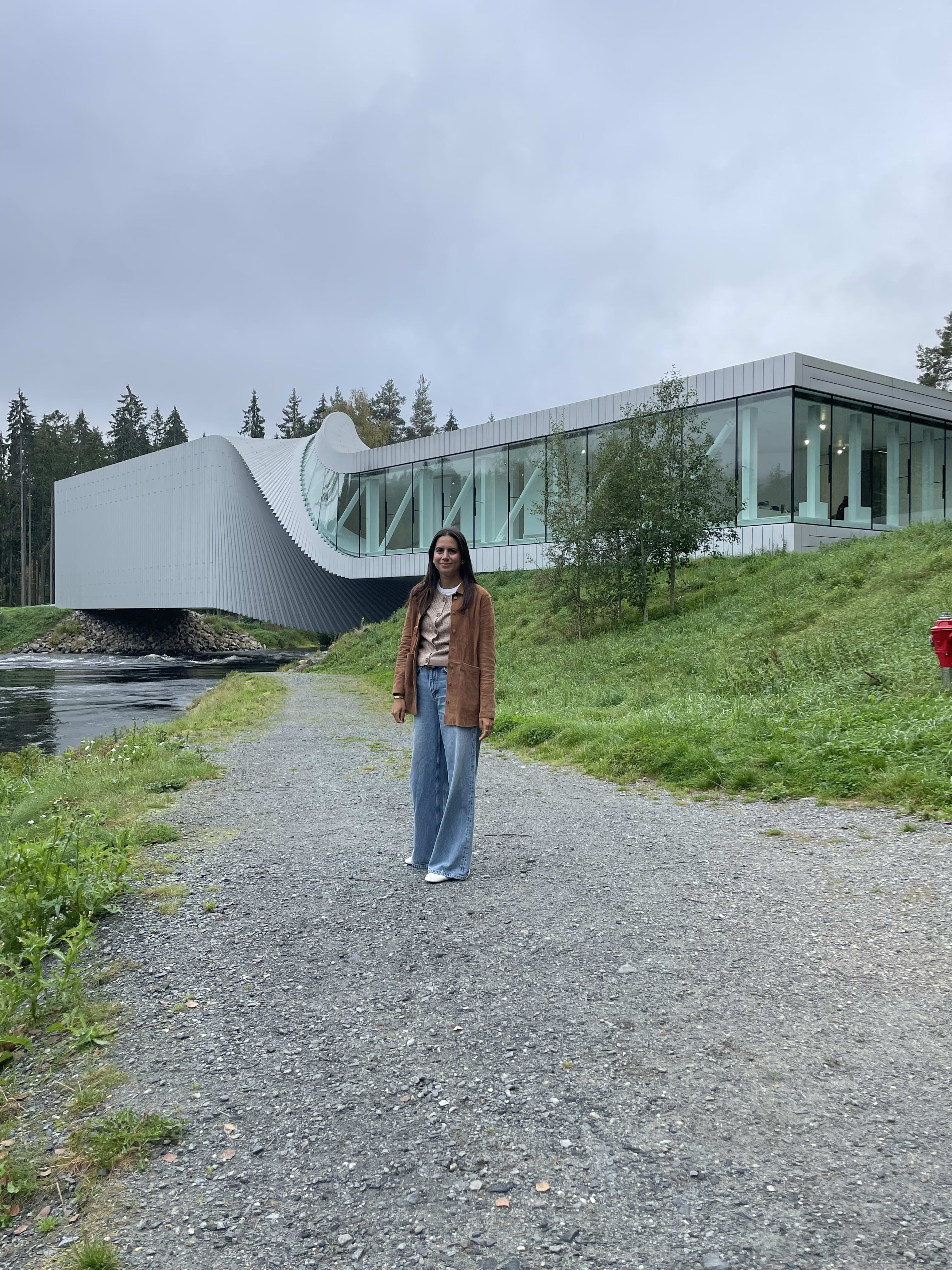 A person stands on a gravel path with a modern building featuring curved architecture in the background.