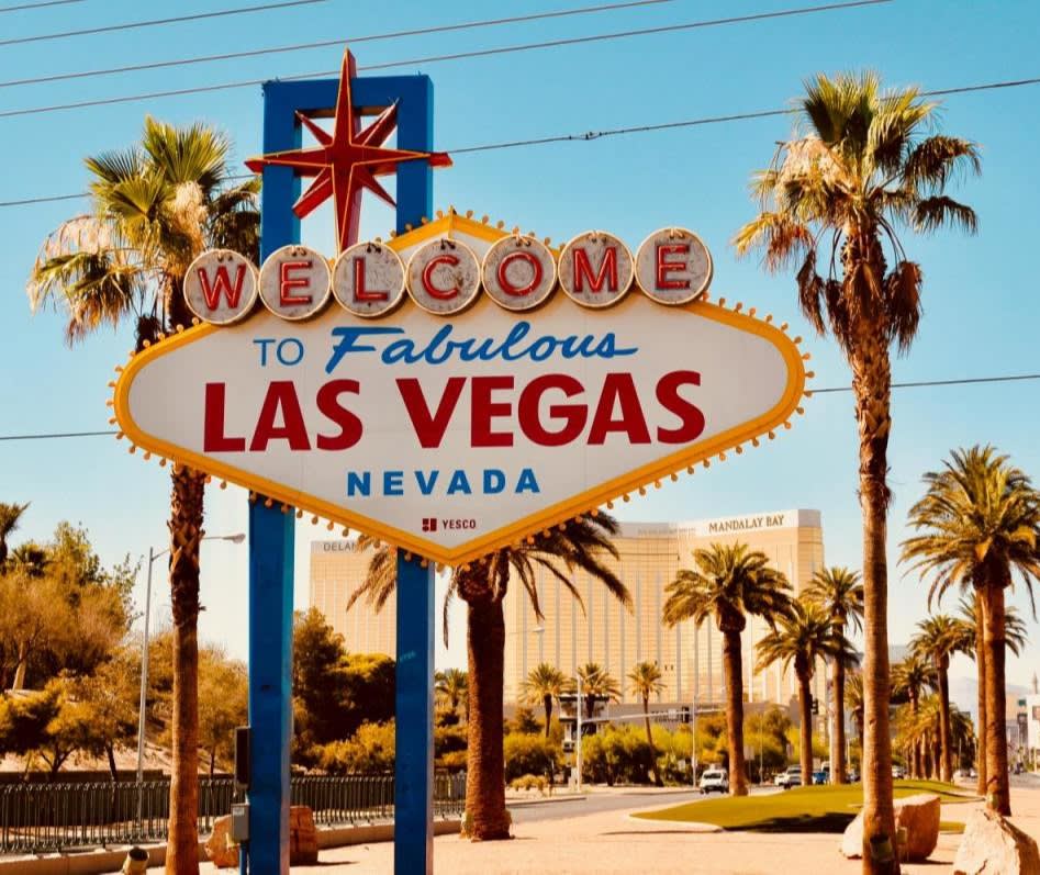 With heavy sun glare: the famous "Welcome to Fabulous Las Vegas" sign standing in front of a palm tree-lined street with Mandalay Bay Casino in the distant background