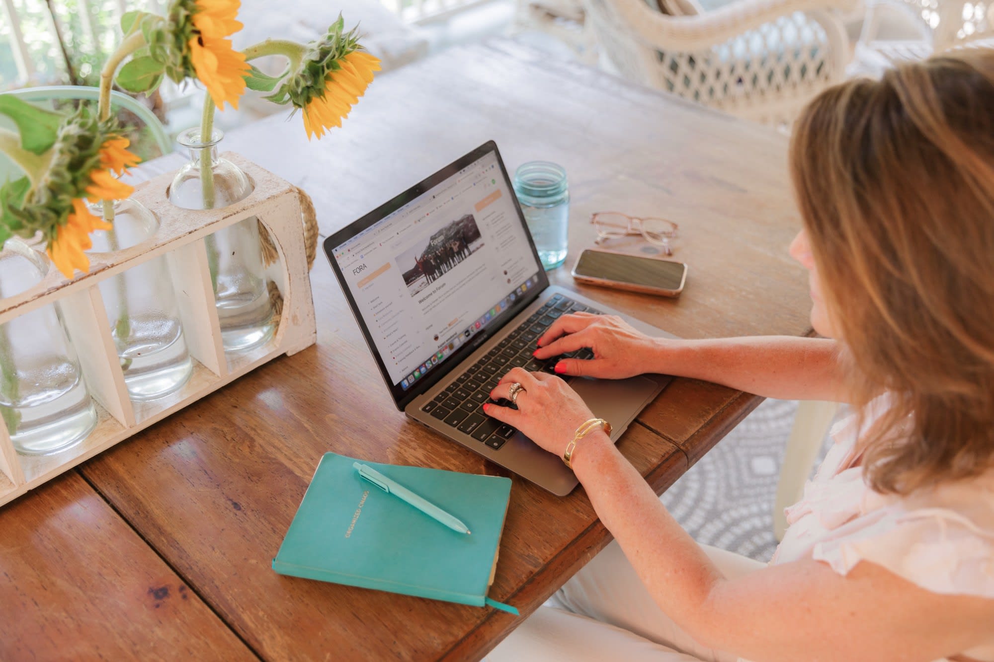 Person working on a laptop at a wooden table with sunflowers, a turquoise notebook, and a smartphone nearby, creating a bright and relaxed workspace atmosphere.