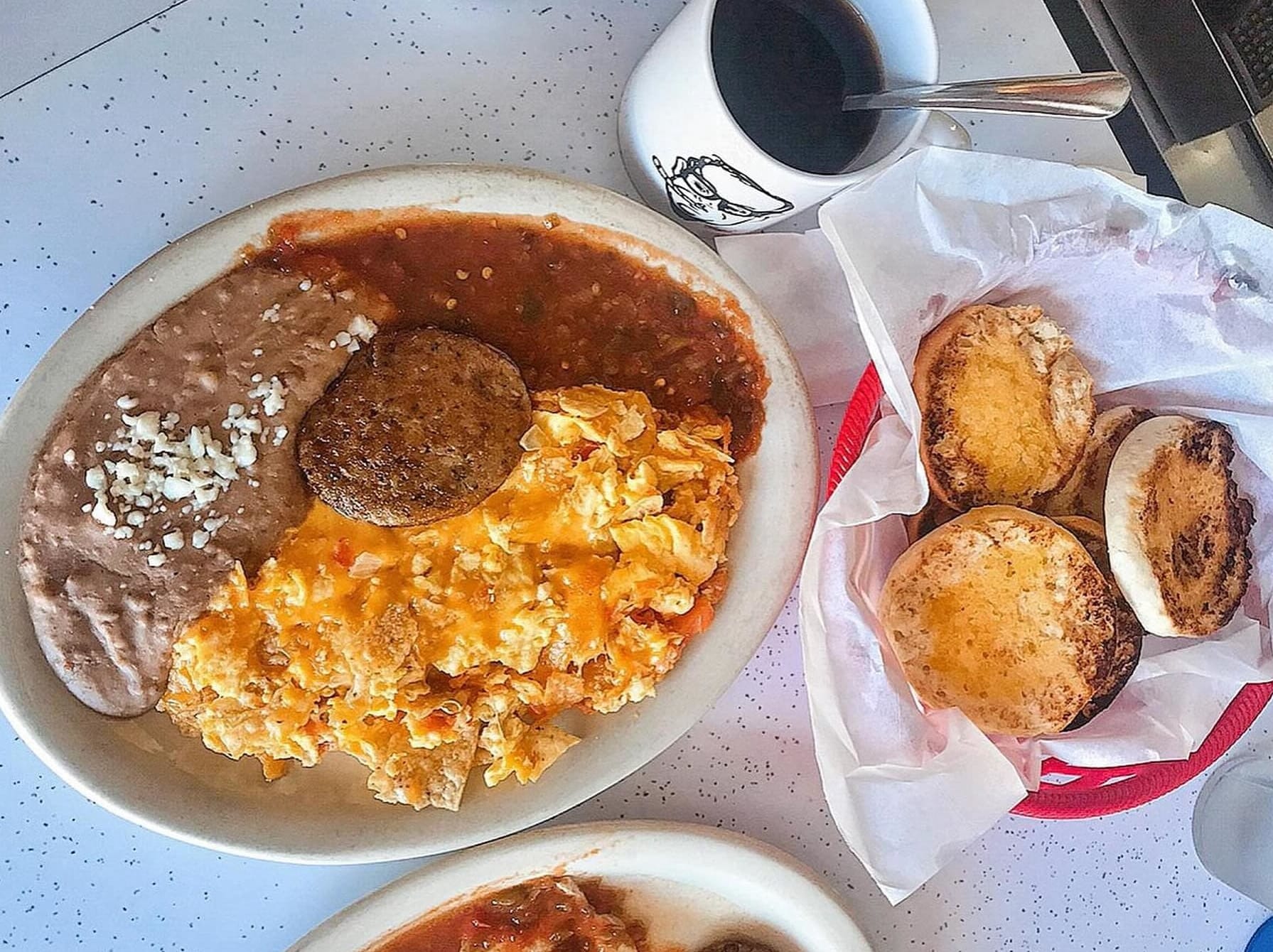 aerial view of a basket of biscuits near a big plate of scrambled eggs, salsa, sausage, and refried beans
