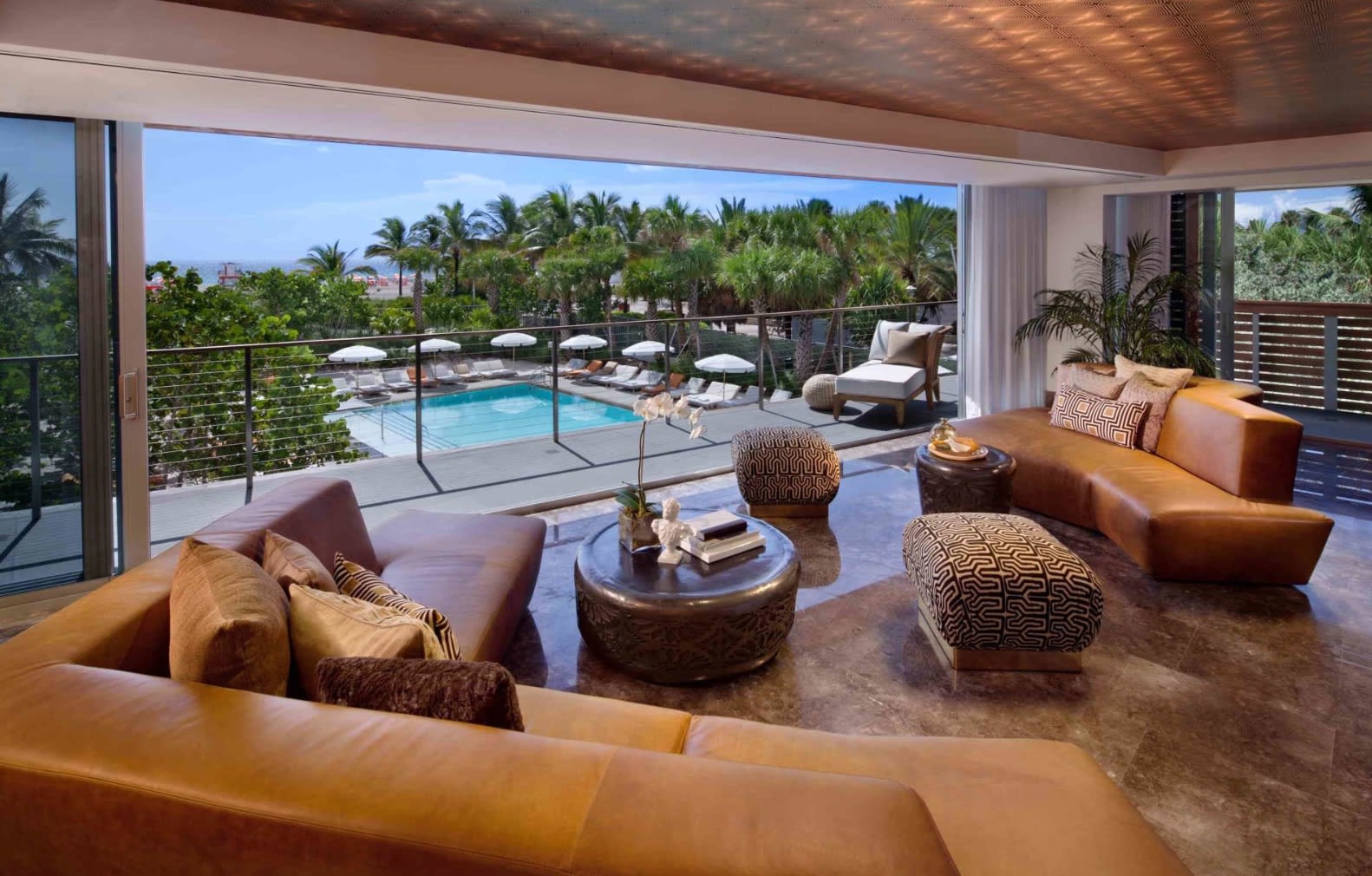 leather couches and round tables overlooking a pool with beach and palm trees in the distance