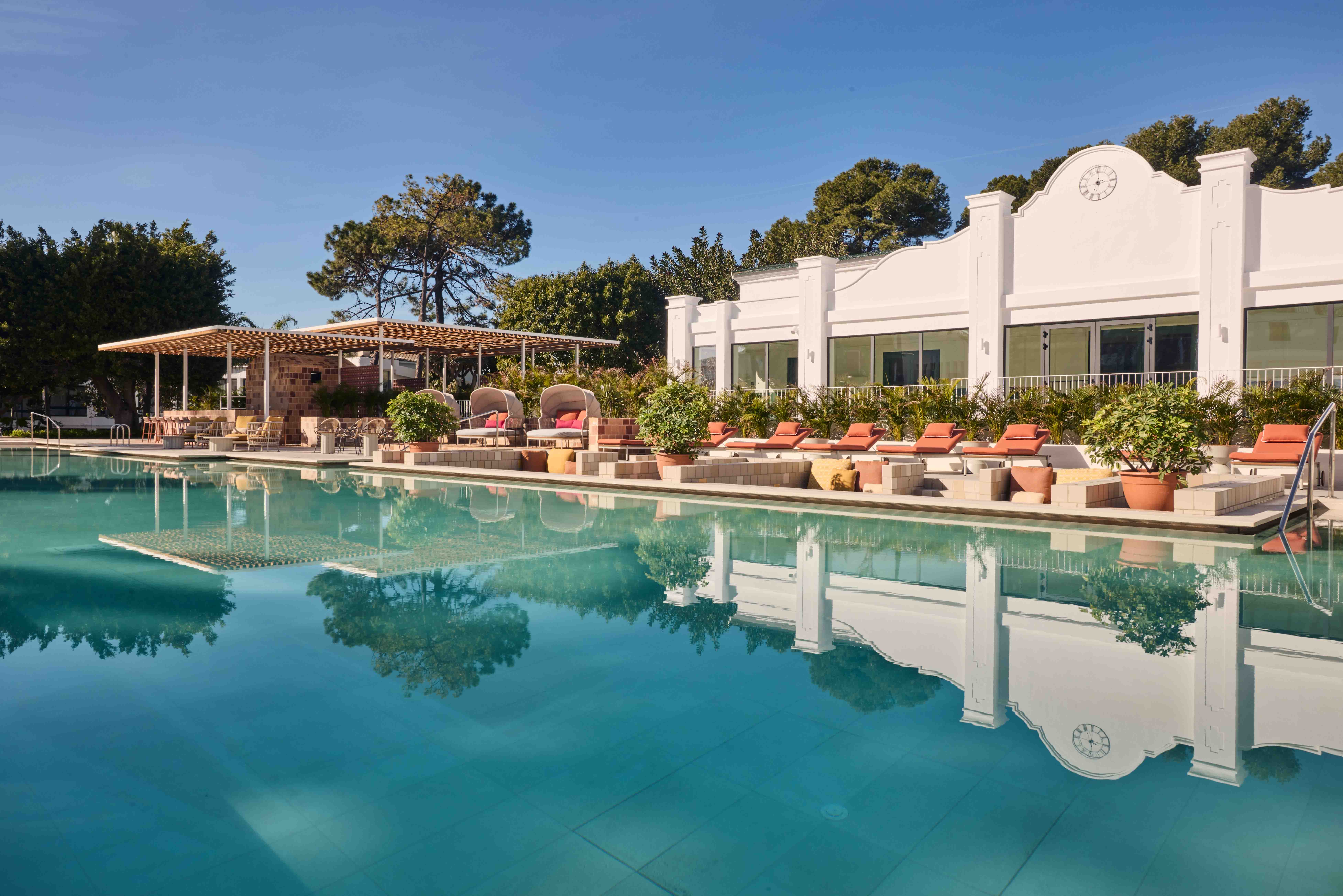 Hotel swimming pool with sun loungers and red cushions on a sunny day with a white building behind
