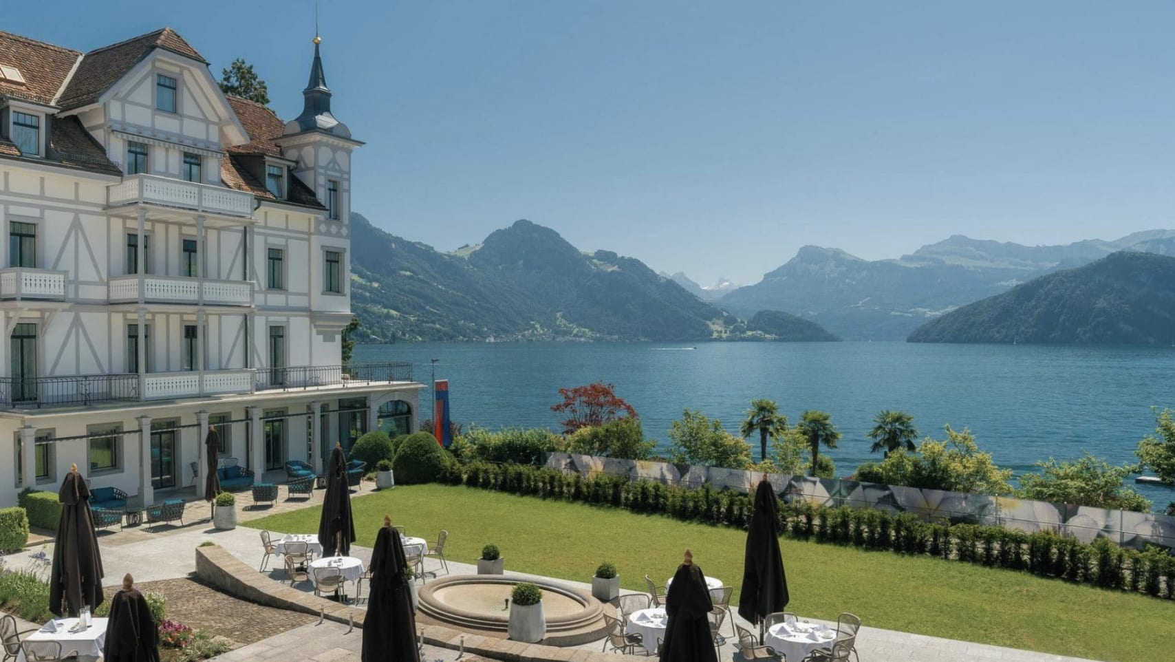 A white hotel building with gardens by a beautiful lake and mountains