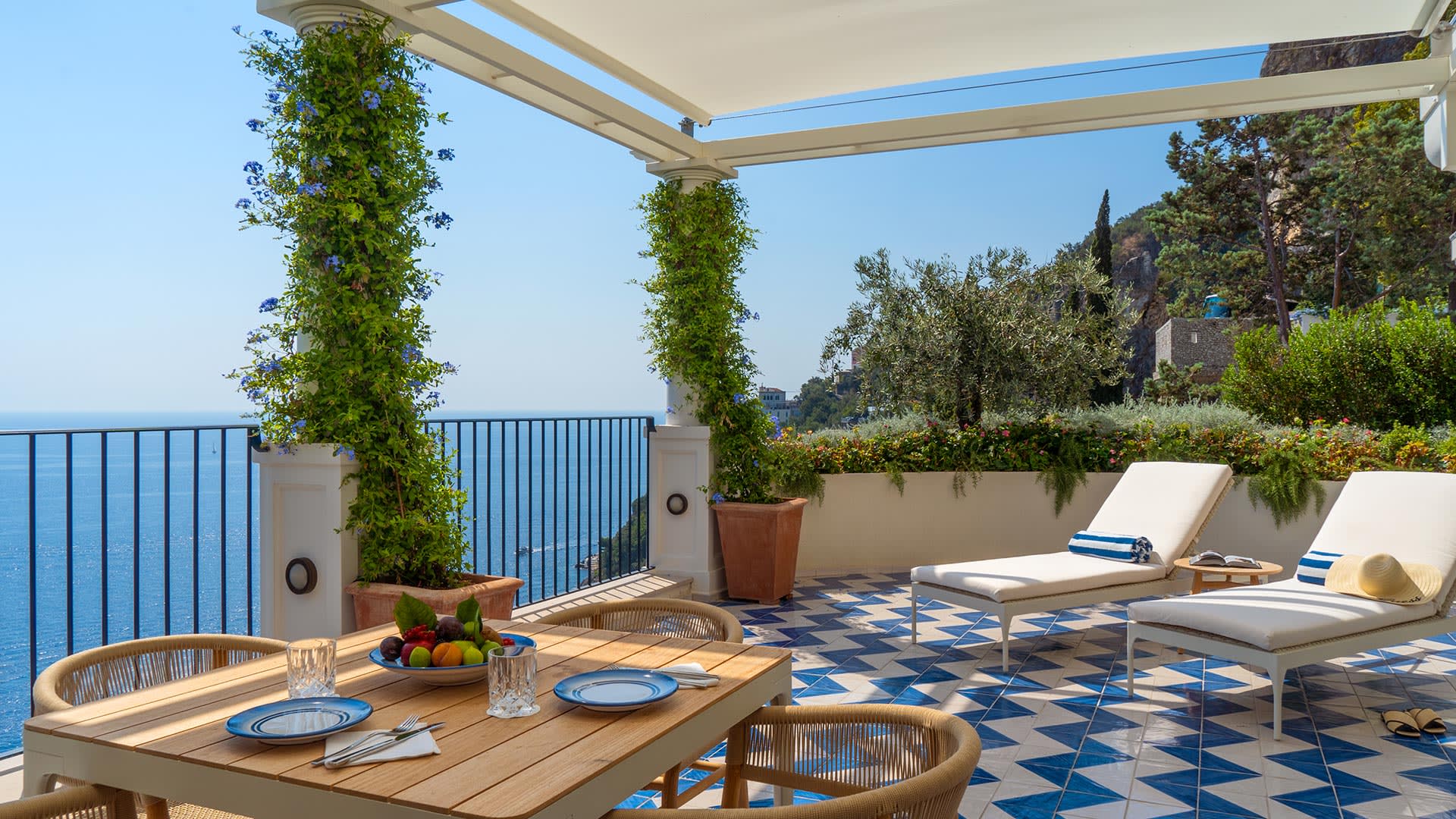 A balcony with blue and white tiled floor with sun loungers and a wooden table and sea views