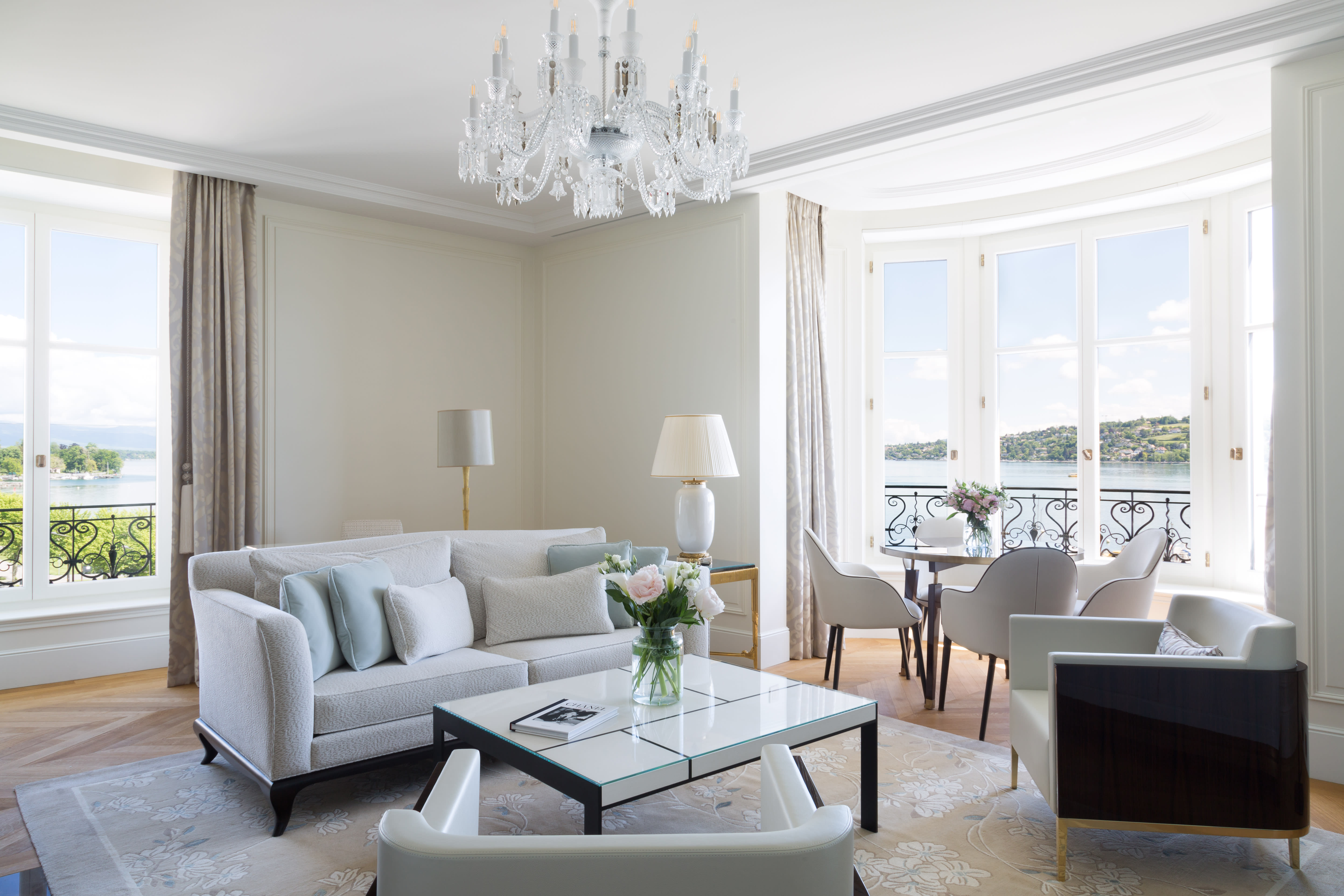 An elegant hotel suite with cream colored furniture, a chandelier and views of a lake