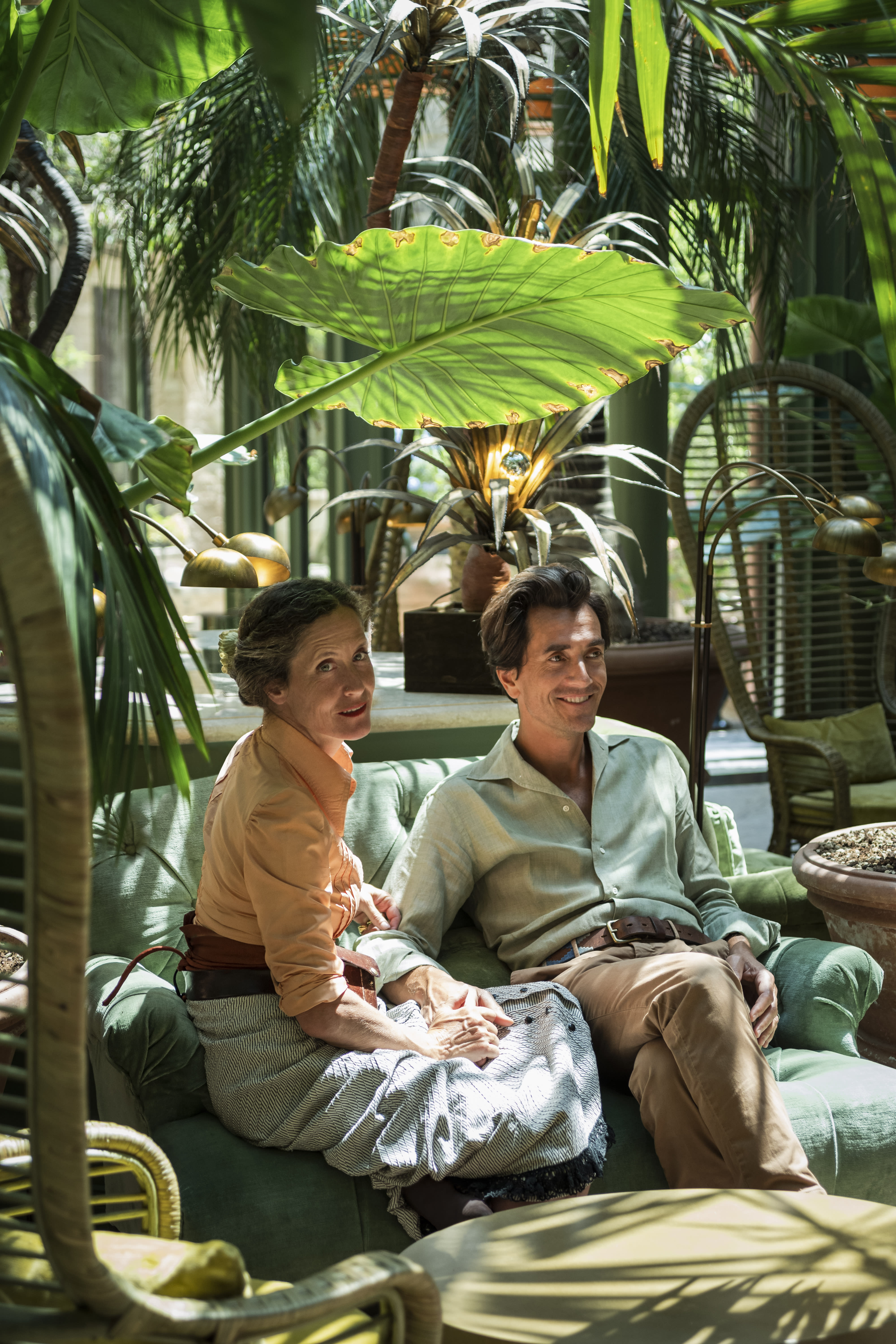 man and woman sitting on a sofa under a green palm tree