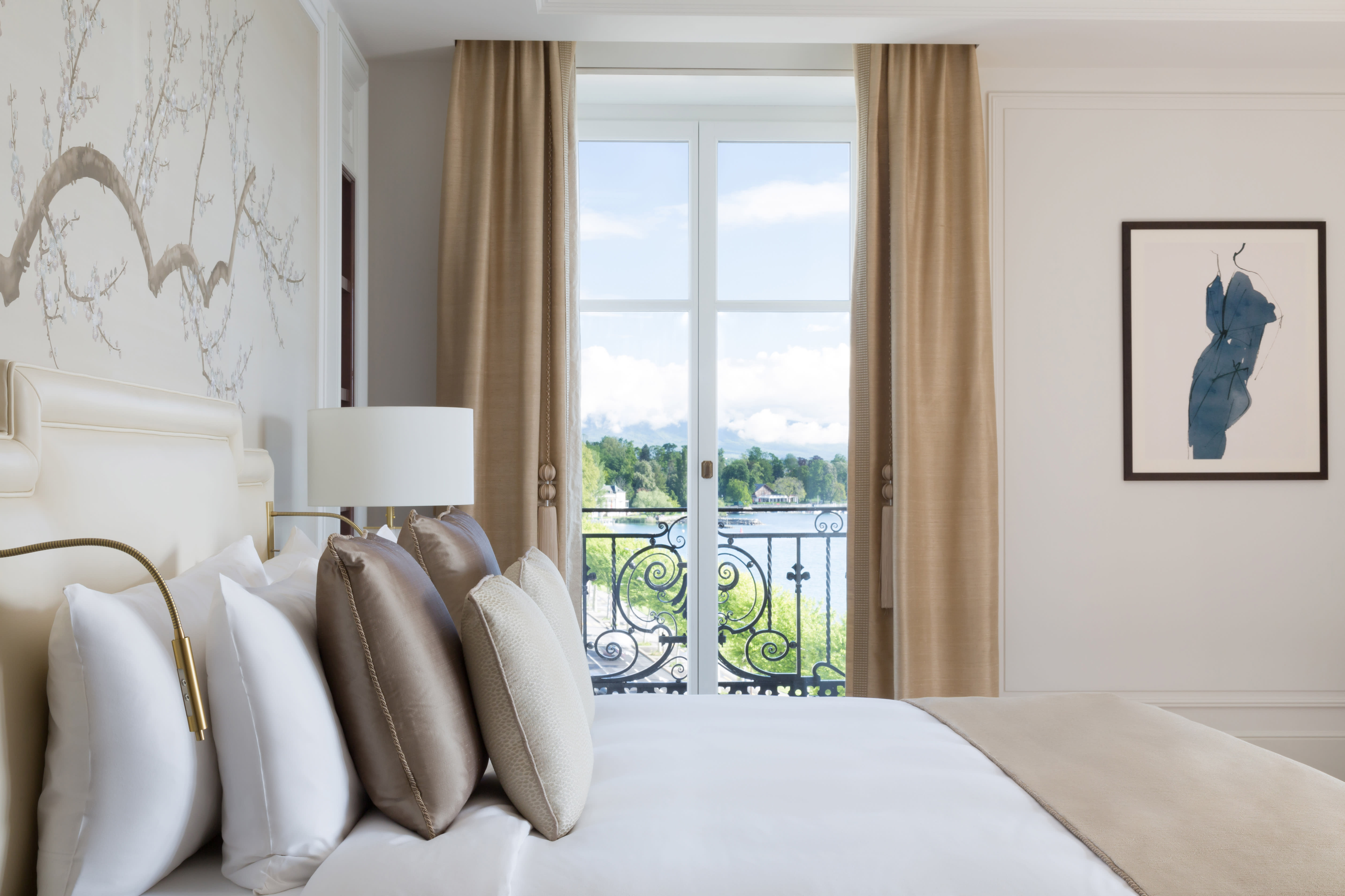 An elegant hotel bedroom with white and cream bedding and a view of a lake through a window