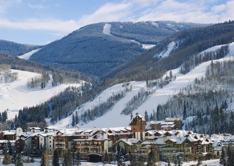 A ski resort in the foreground with a landscape view of a snow covered mountain in the background with ski slopes cutting through forest
