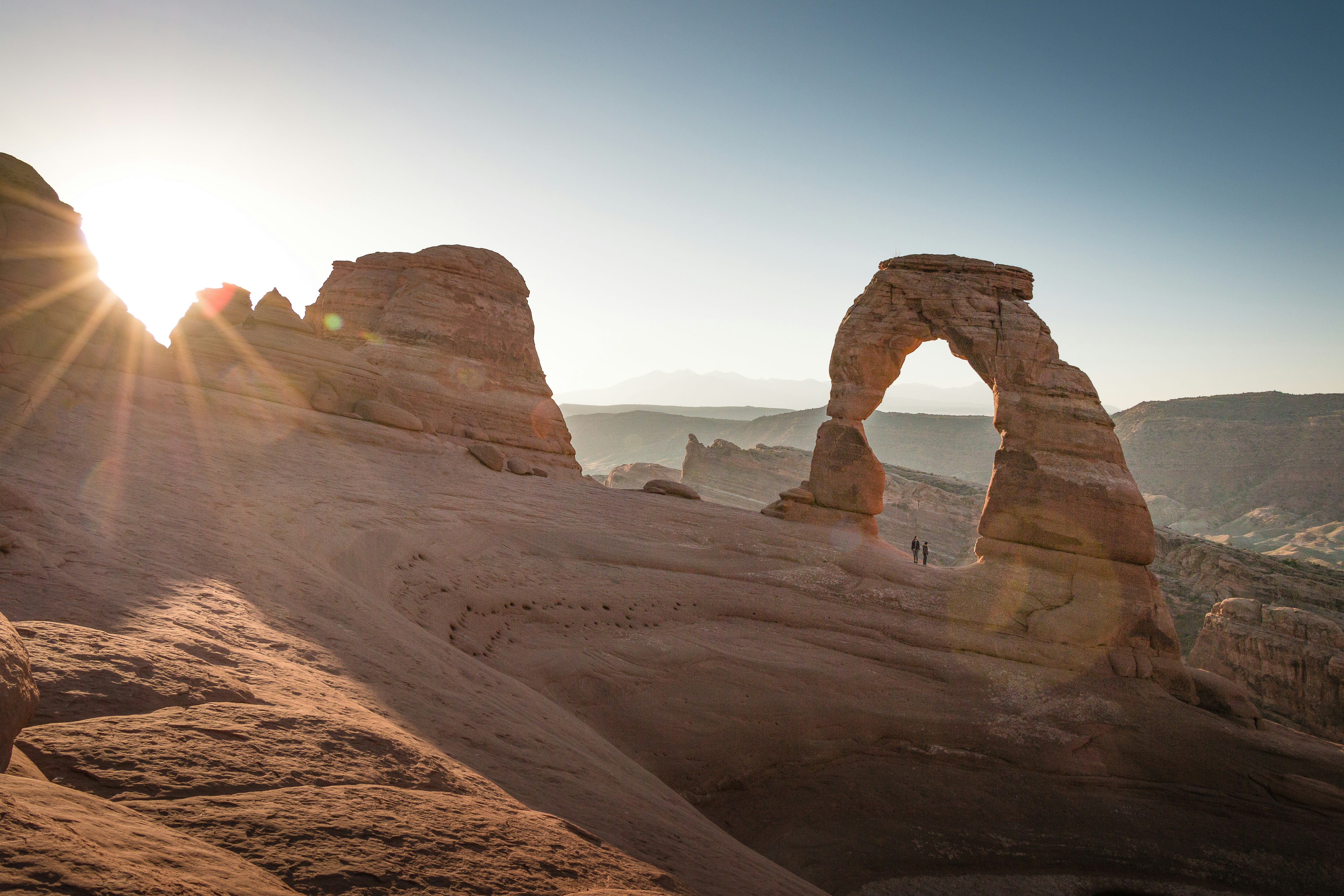 A landscape view of a desert landscape with tan rocks and an arched rock with the sun shining just above the top of the rocks