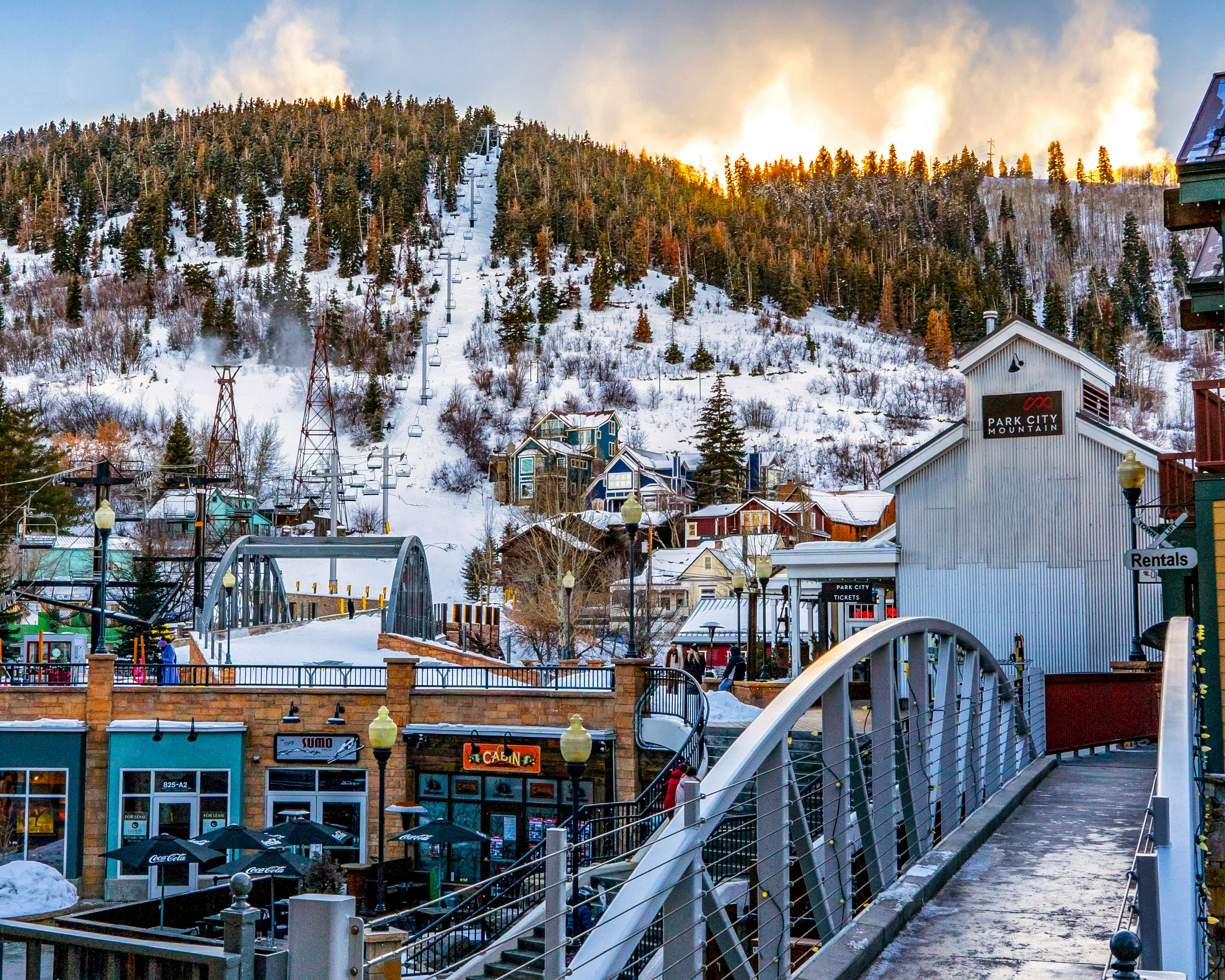 A mountain town with an empty pedestrian bridge leading to brick shop buildings below a snow covered mountain with a ski lift and trees at daytime
