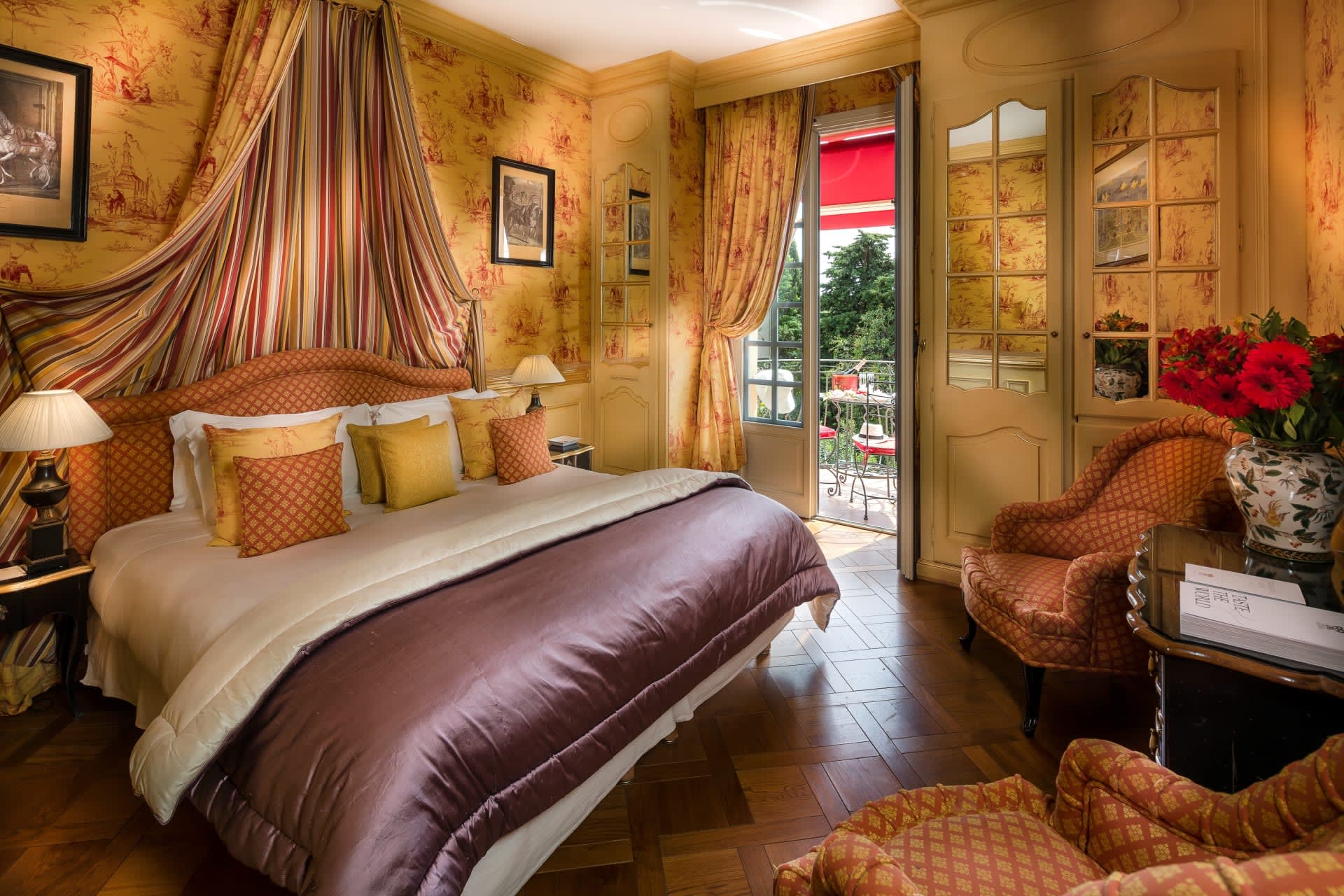 A classic French styled bedroom with yellow patterned wallpaper, orange pillows and chairs, purple comforter and wood floors