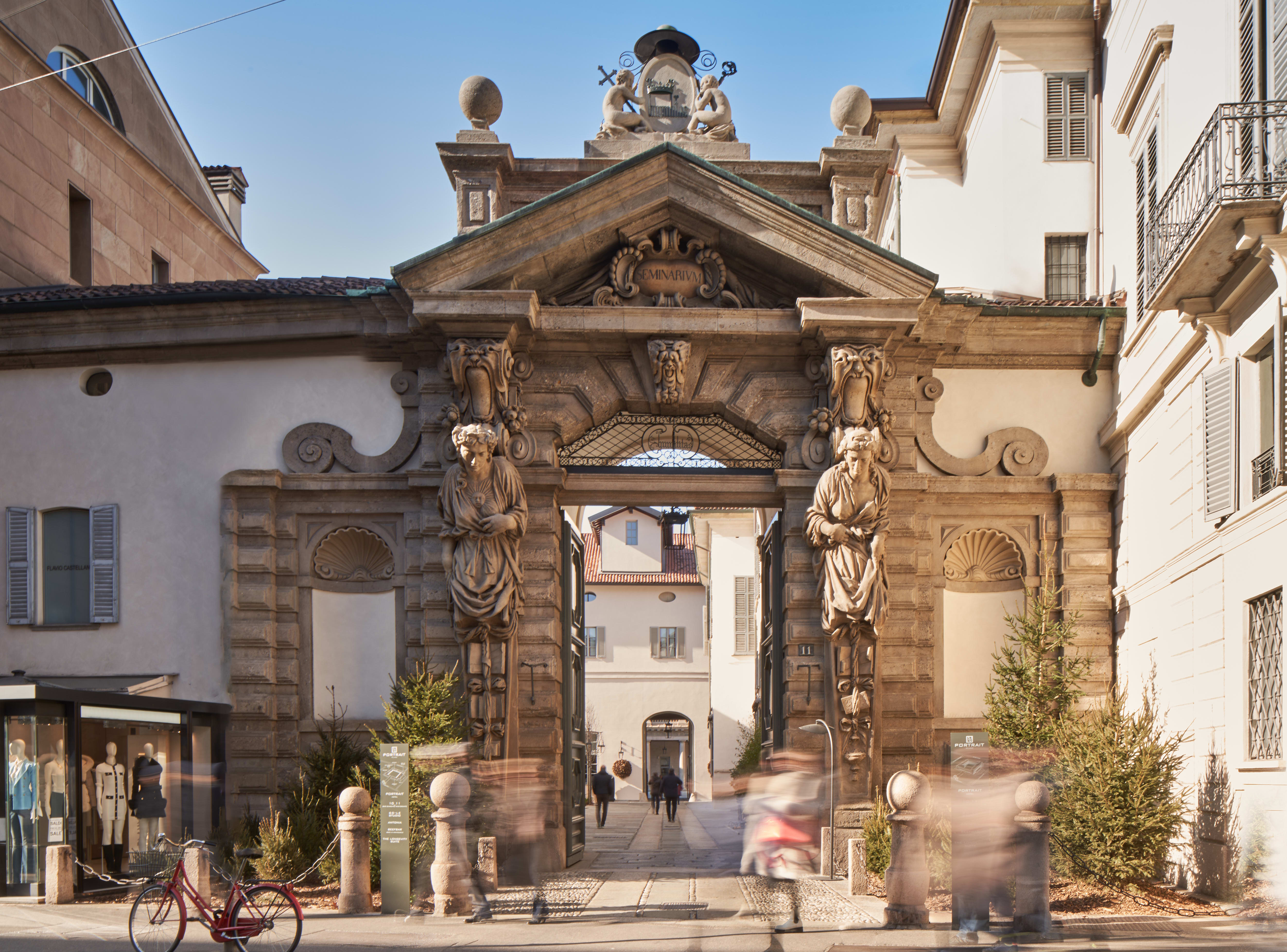 The arched, stone entrance of a piazza in Milan with ornate stone carvings in brown and white.