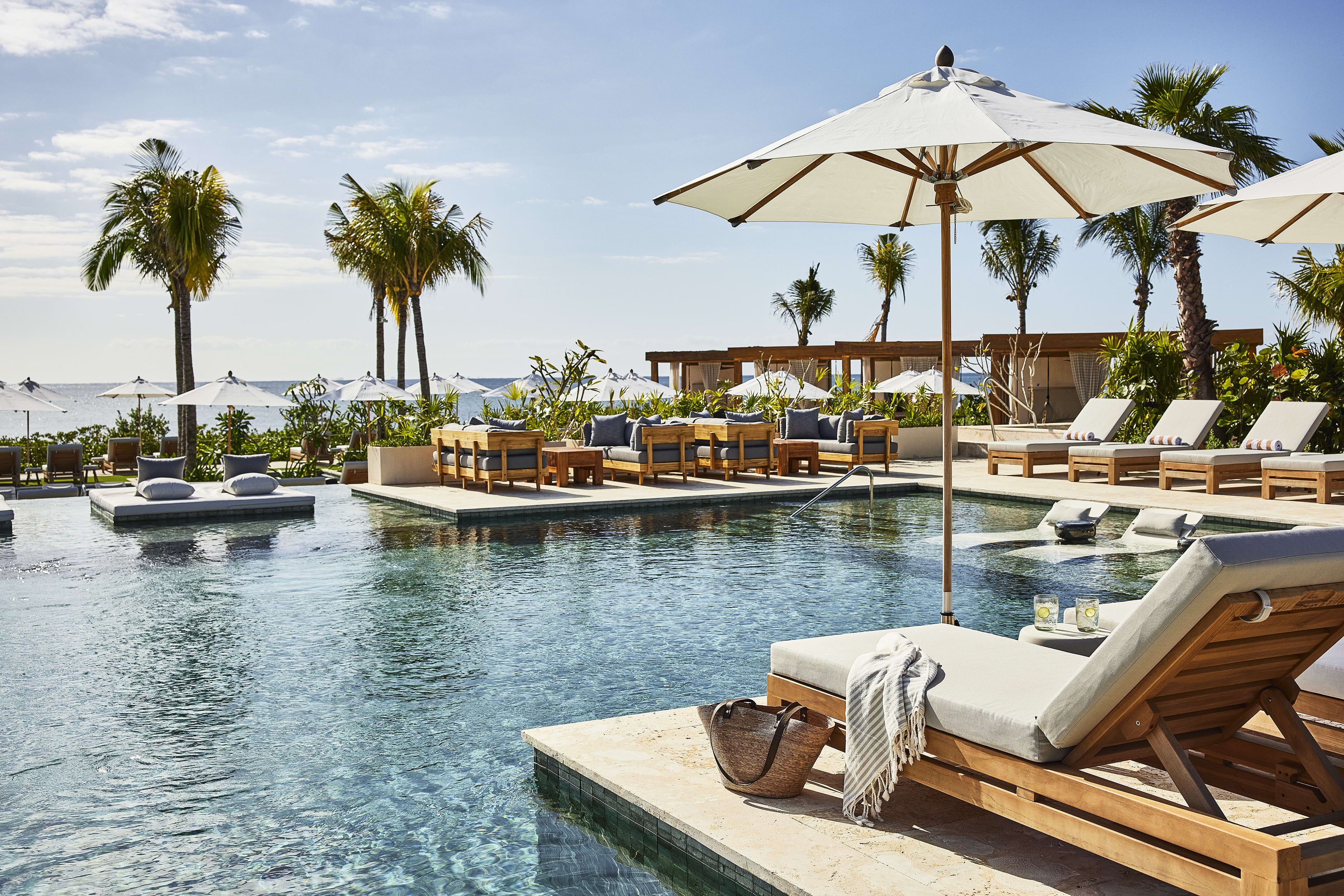 A hotel pool area with plush cream loungers, umbrellas and palm trees