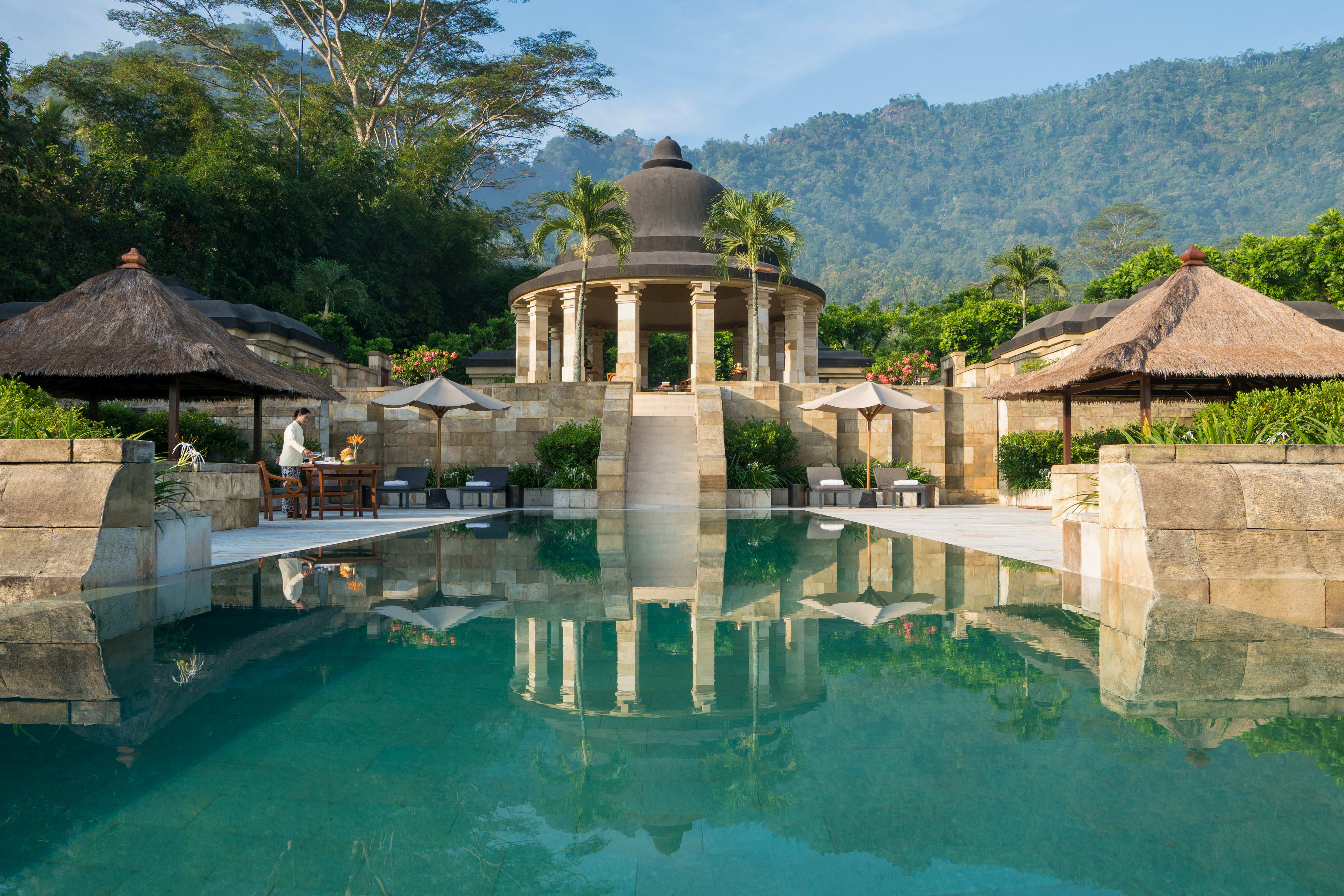 A hotel pool area with a traditional temple-style building and lush greenery and mountains in the background