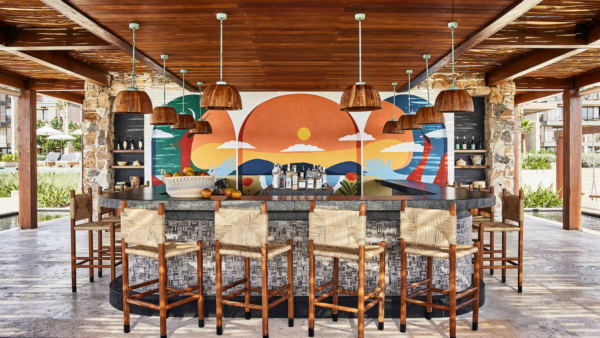 A colorful and modern bar area of a hotel with wooden bar stools and hanging lamps with a mural.