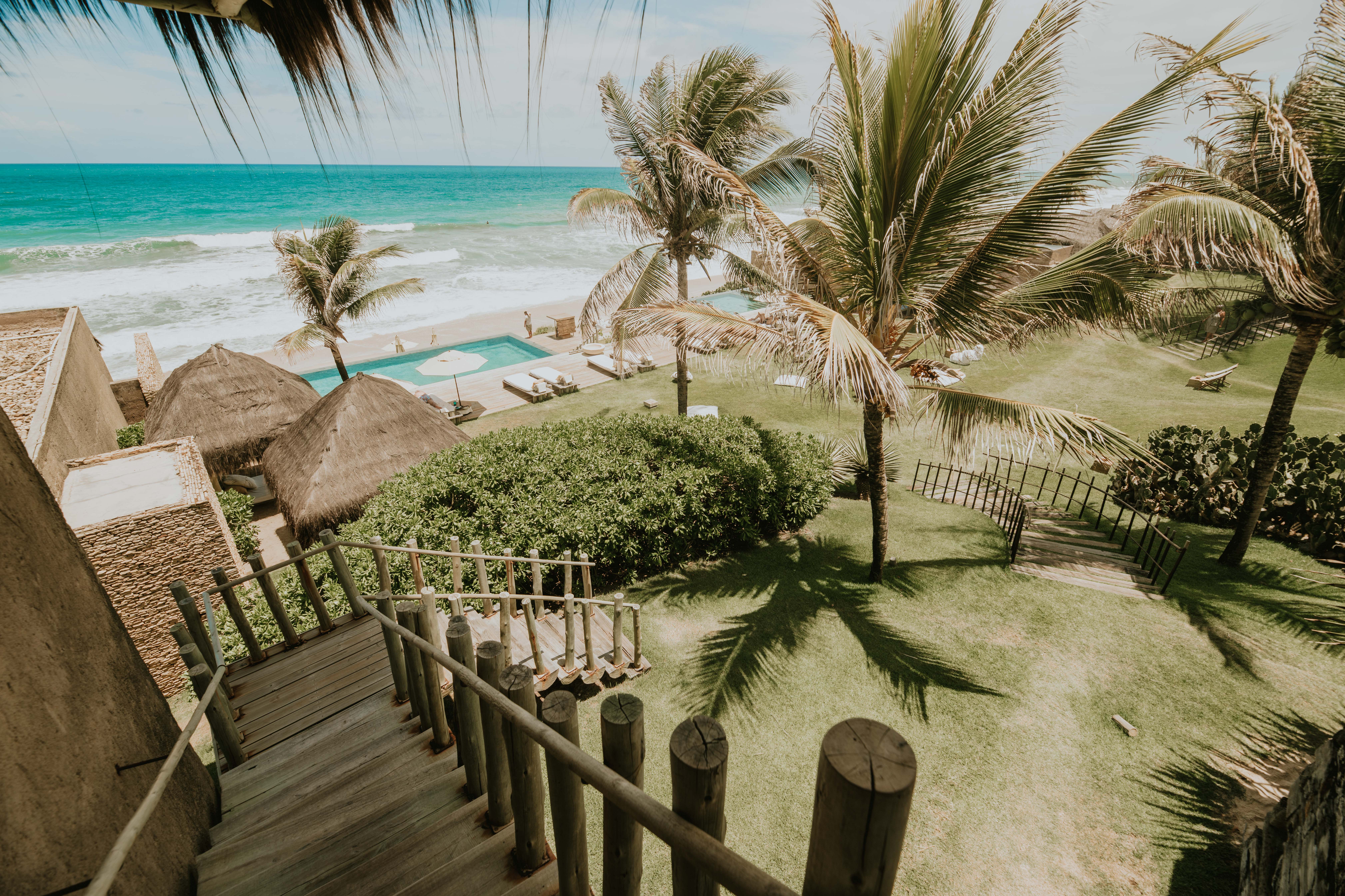 A view down wooden stairs towards a beach with swaying palm trees and green areas