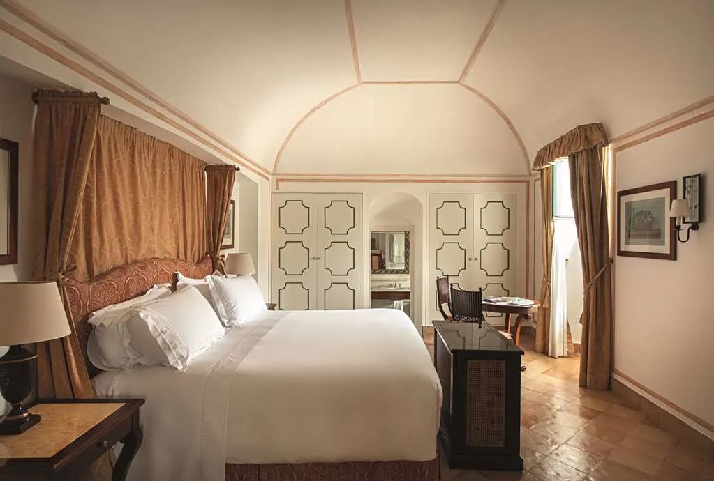 Side view of a hotel room with tan curtains behind the white bed, rounded ceiling and contemporary decor