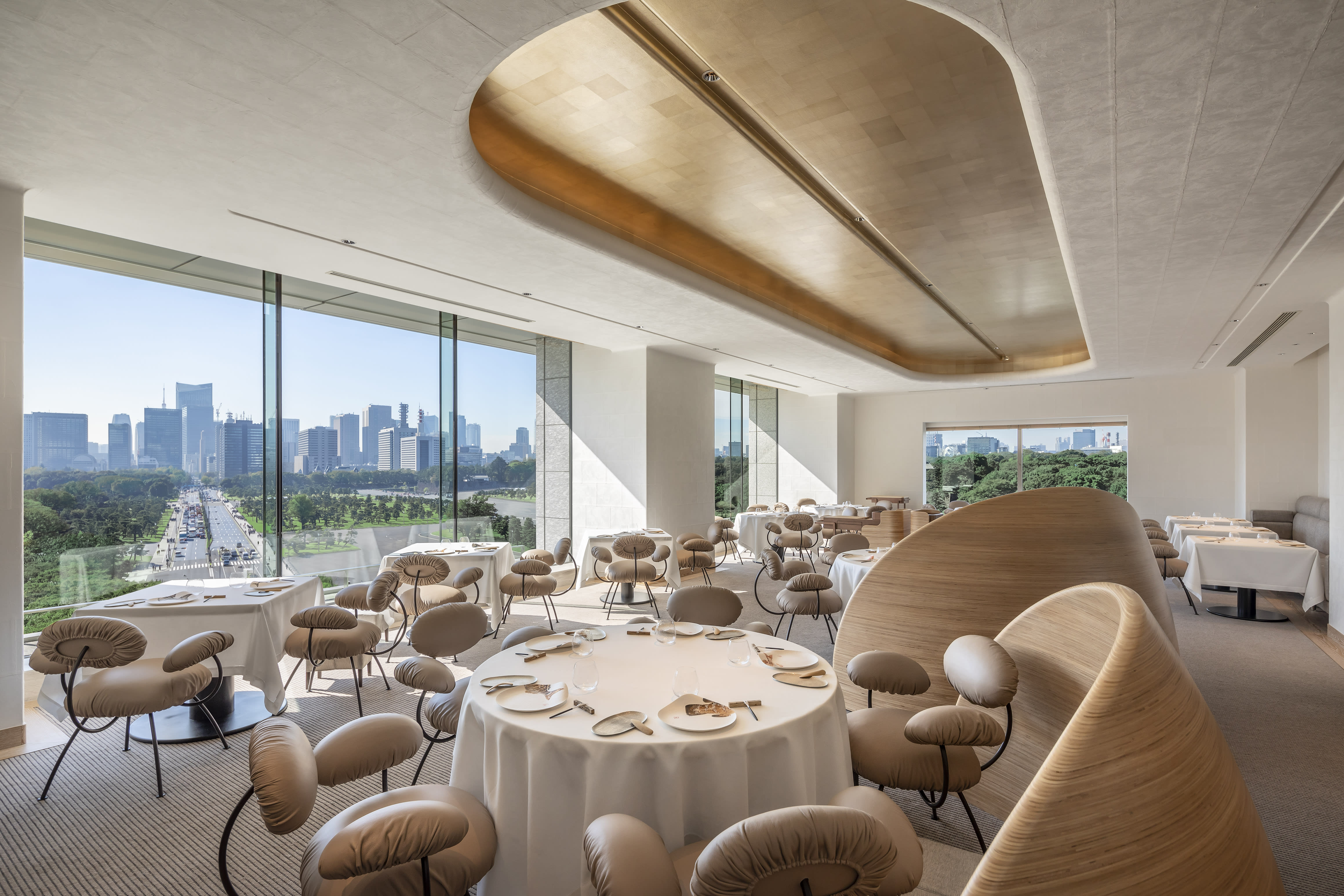 A bright, modern hotel dining space with gold chairs and ceiling decoration with floor-to-ceiling windows showing cityscape