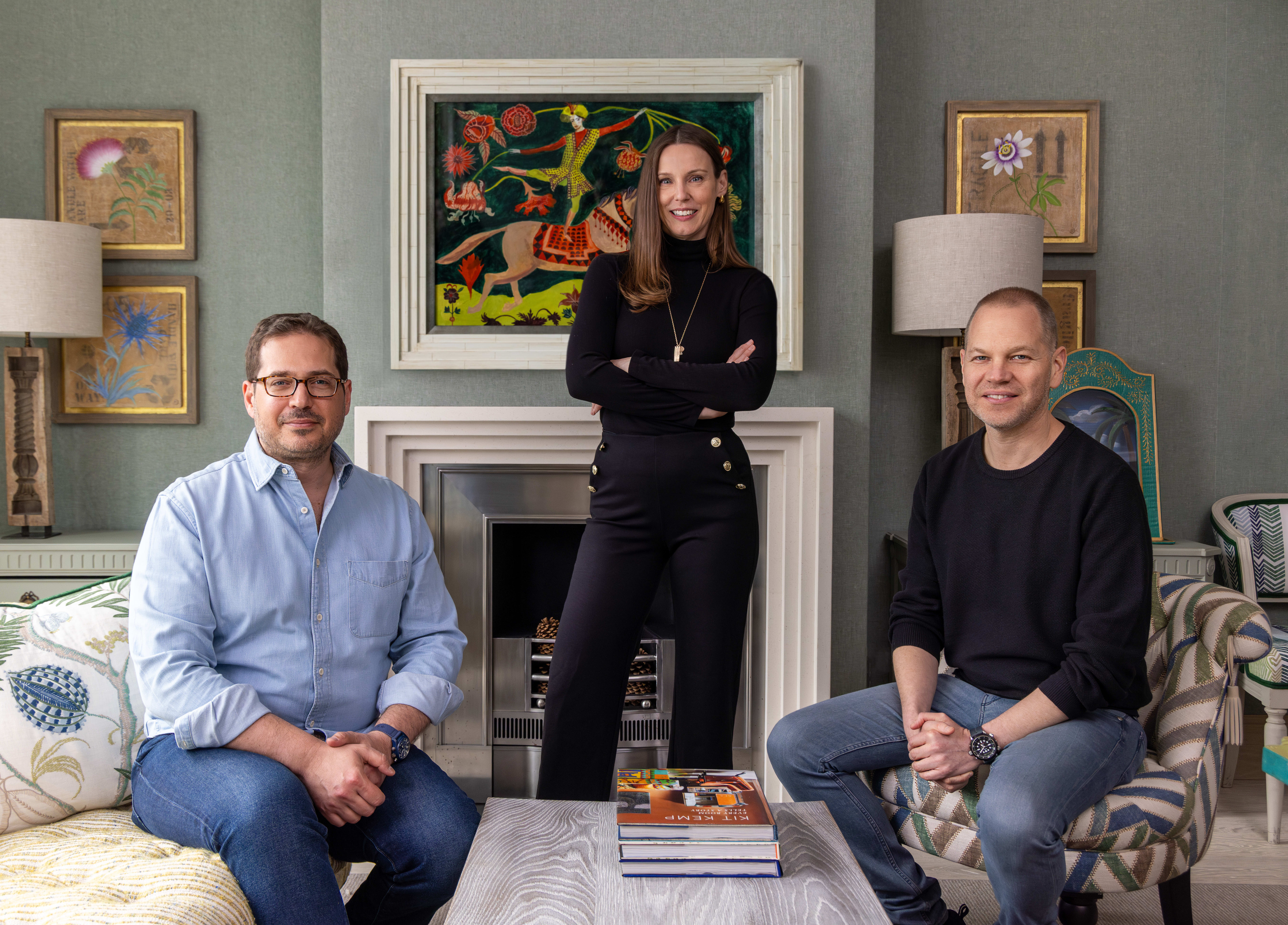 two men sitting in a living room and a woman standing with arms crossed in between