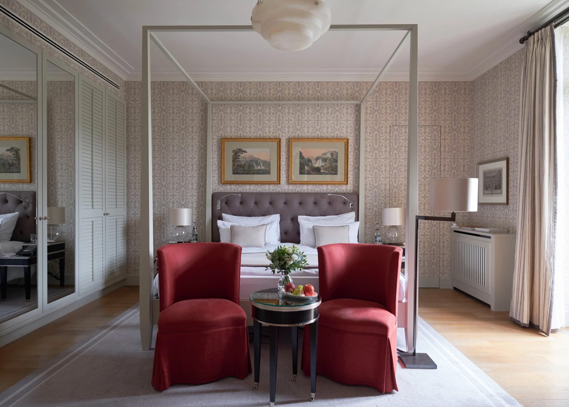 A hotel room with muted wallpaper walls, two red chairs in front of a modern four poster framed bed