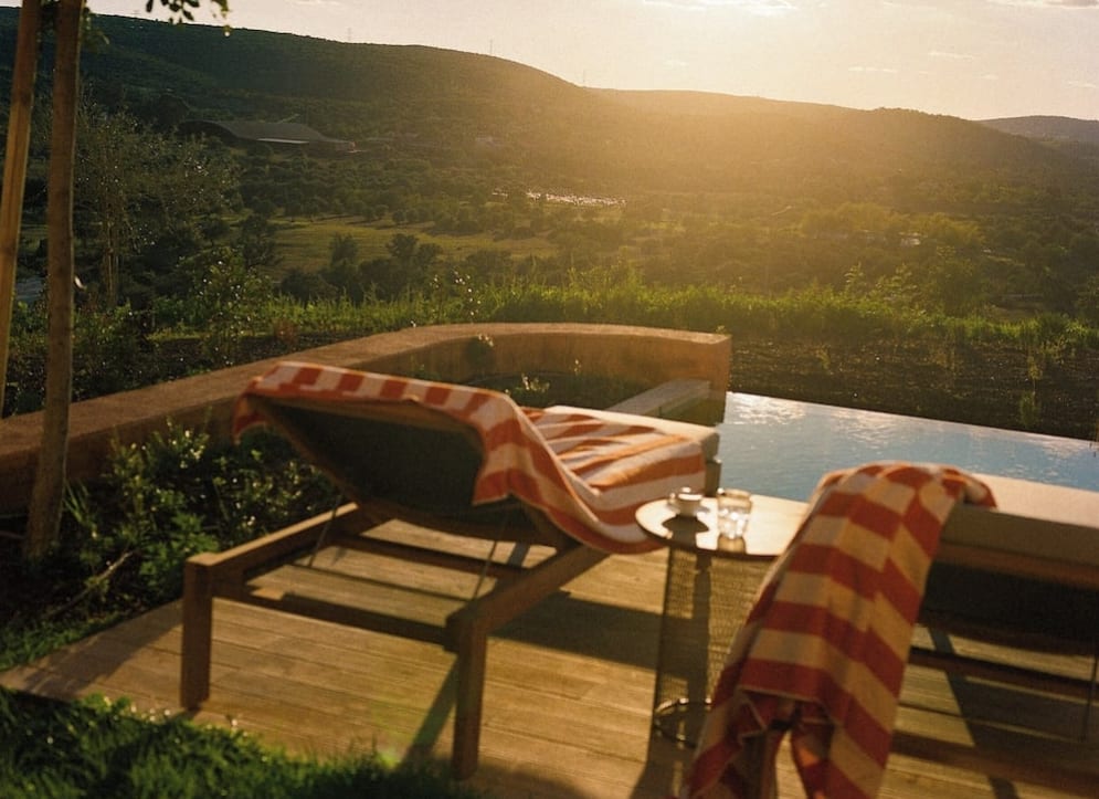 Two sun loungers on a deck with red and white striped towels and a small pool overlooking green landscape with the sun low in the sky.
