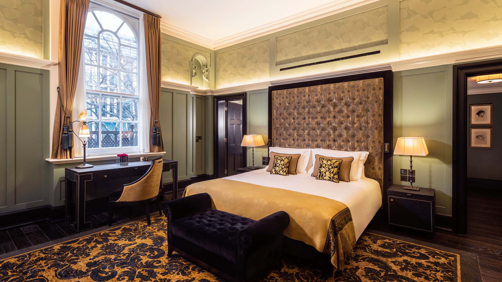 A contemporary hotel room with dark brown & tan patterned carpet, sage colored walls, and light maroon fabric headboard with a window, desk and chair next to the bed
