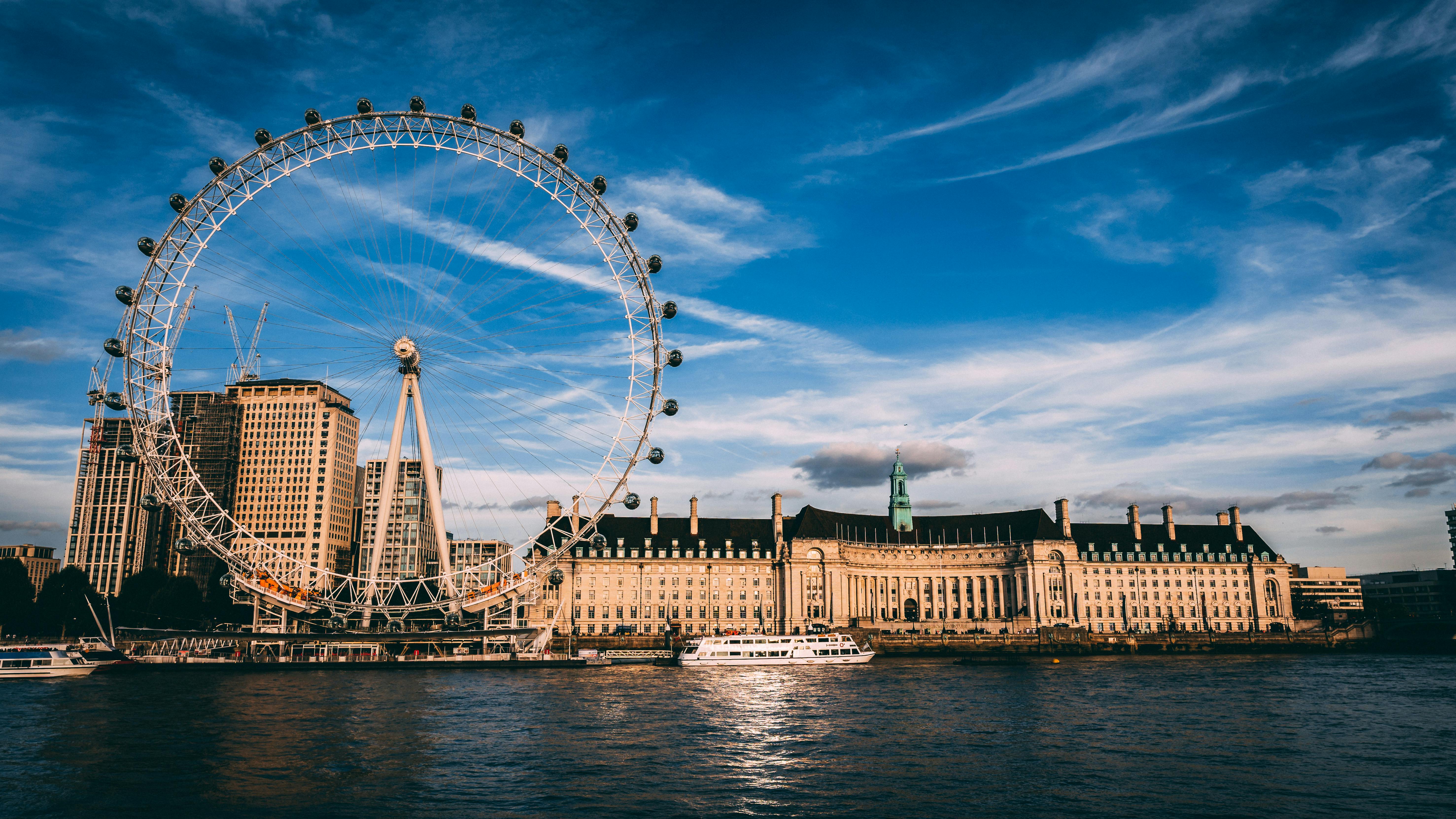 River in the foreground with historic buildings and a giant ferris wheel on the other bank during day with a blue sky
