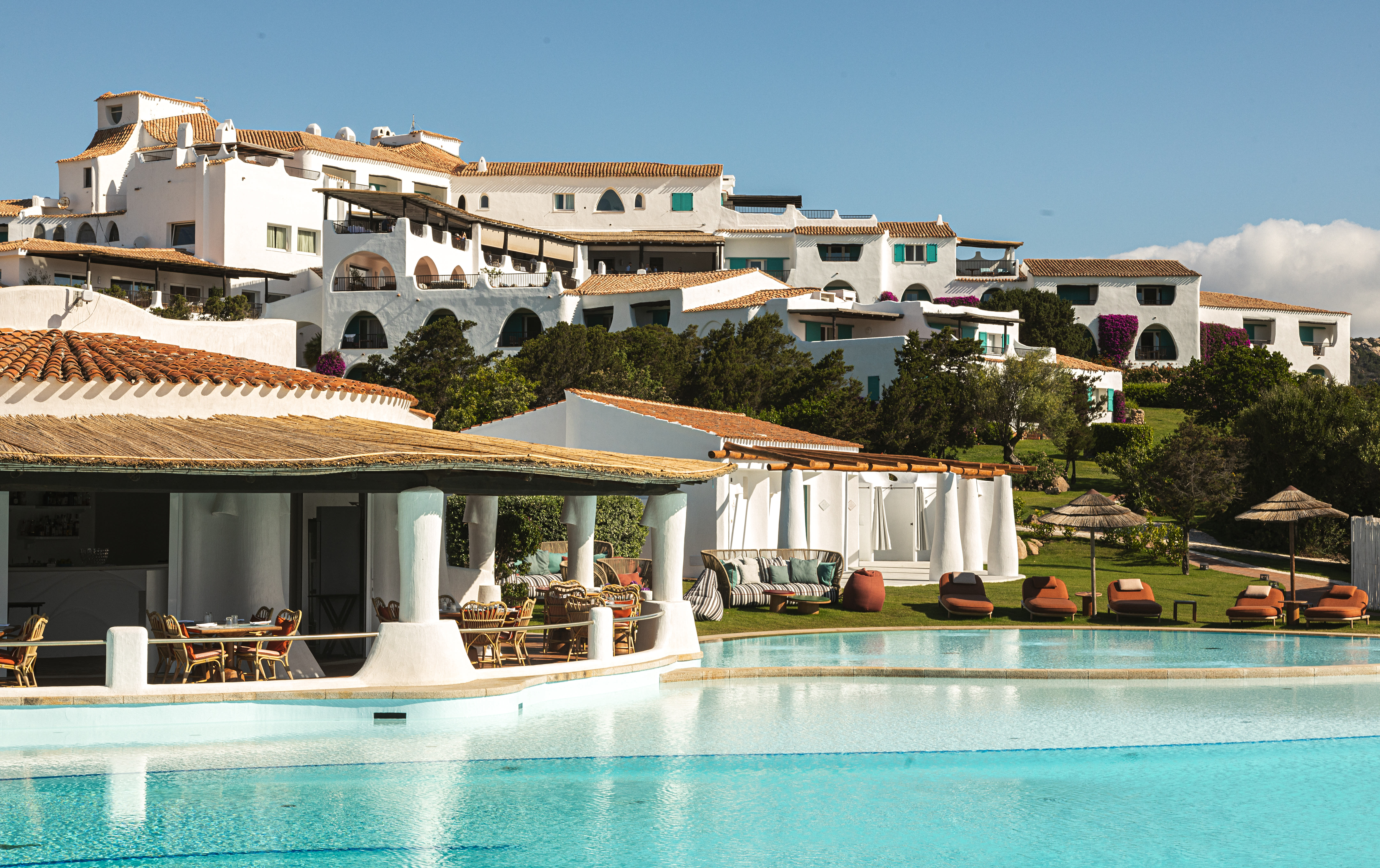 A hotel pool area with cabanas, a restaurant and whitewashed buildings on a hillside behind