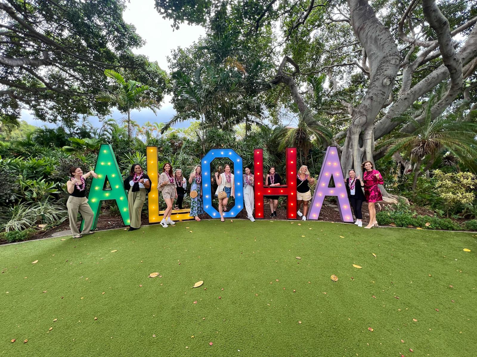 a group of women stand amongst colorful letters that say "Aloha."