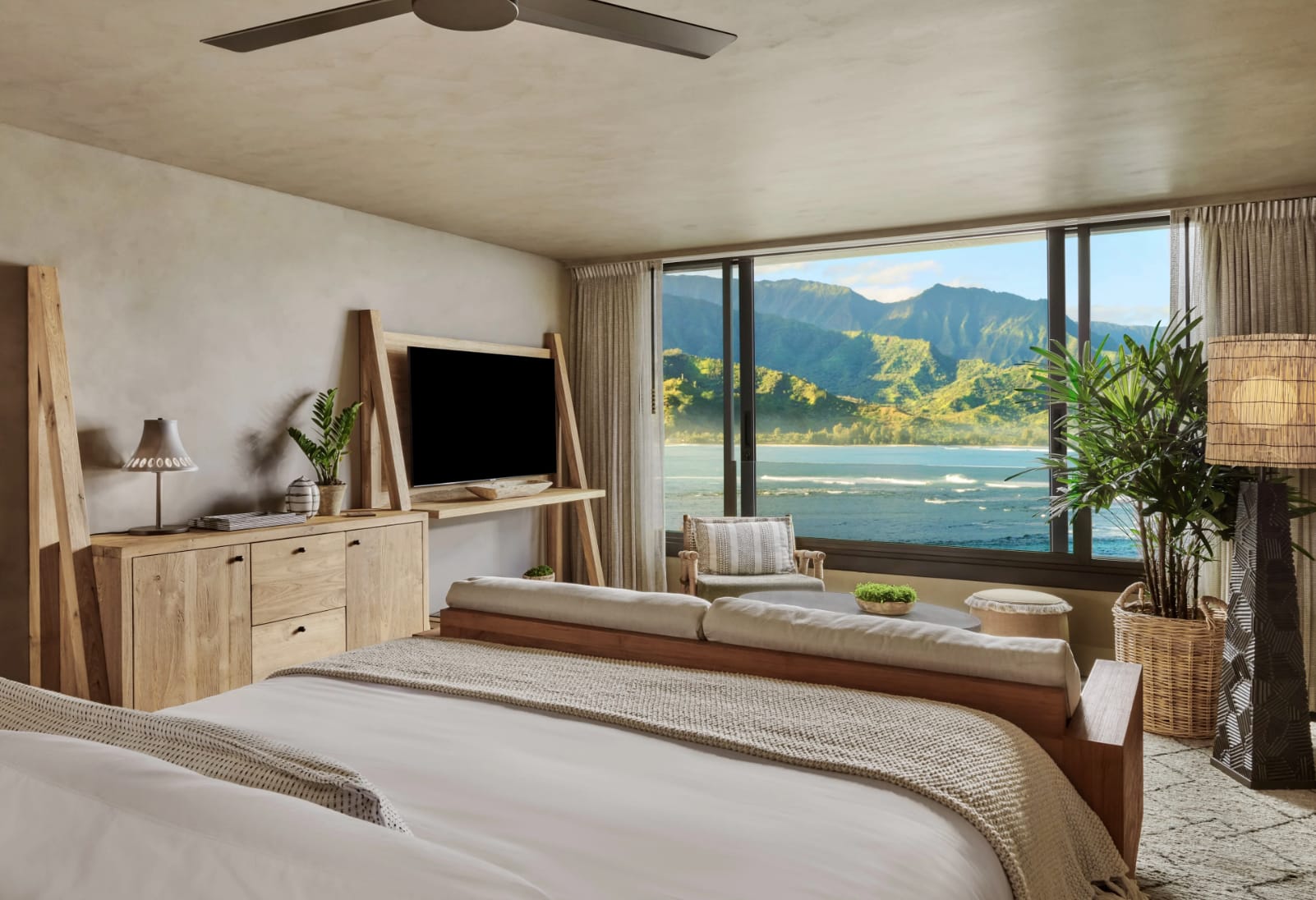 A neutral colored room with bed facing wall-to-wall windows with tropical mountains and ocean in the distance