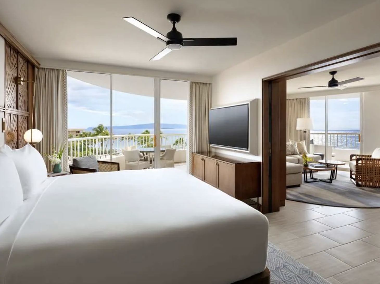Expansive hotel room with bed to the left facing a TV and under a fan with separate seating living room off to the right, both with sliding glass doors and ocean beyond