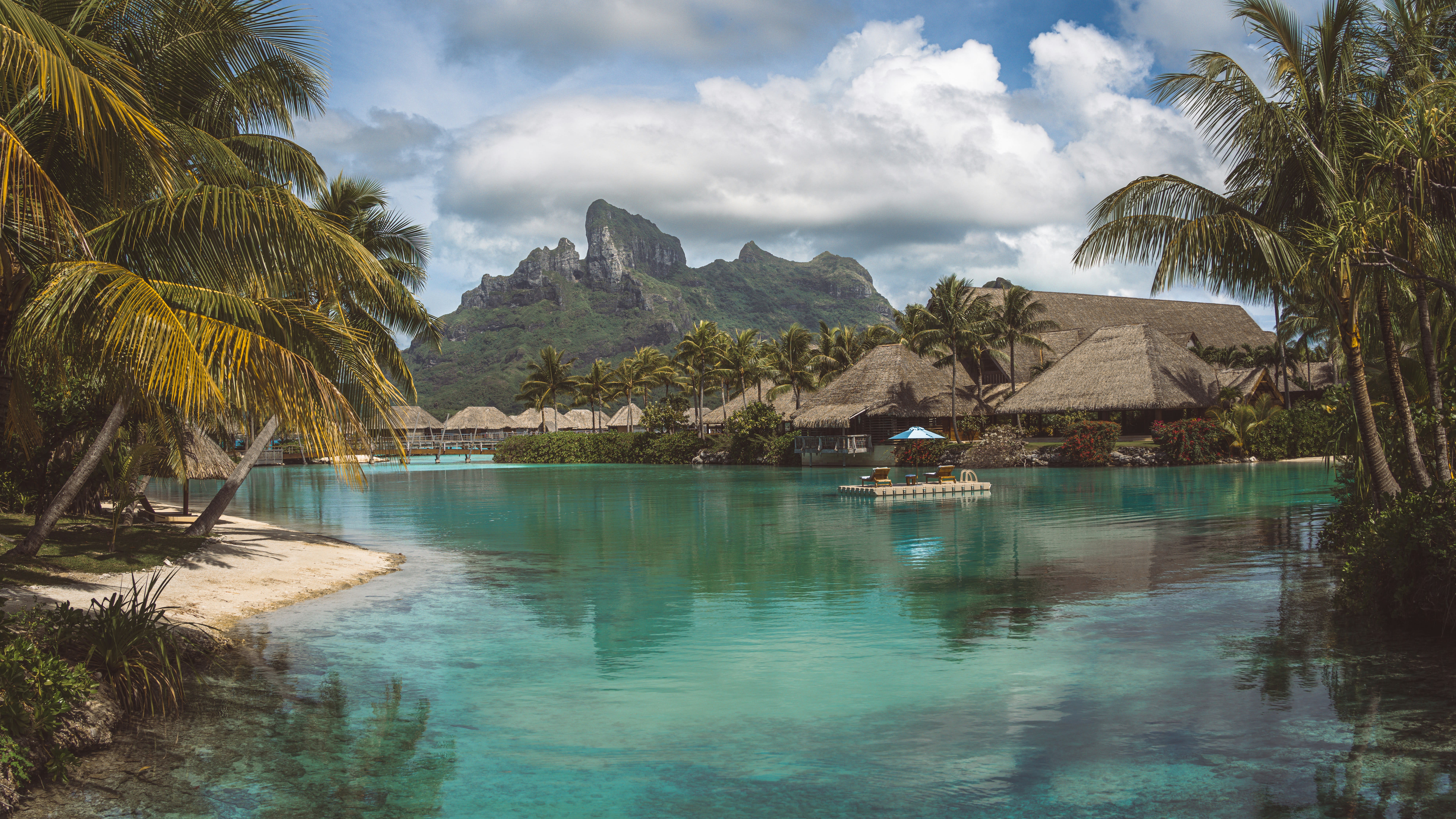 A tropical lagoon surrounded by palm trees and the Four Seasons Resort Bora Bora, with mountains in the distance during day