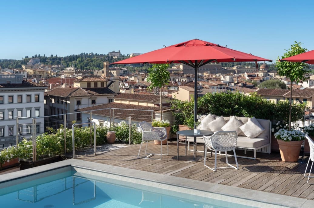 A hotel terrace with white seating and a red umbrella and pool with city views