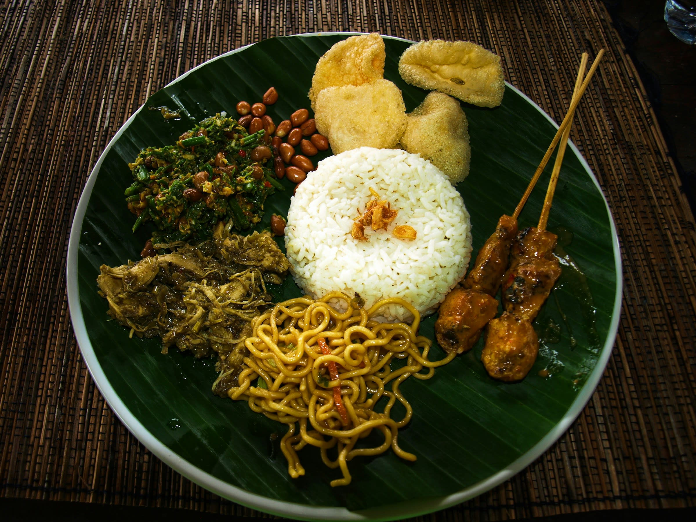 A plate with satay, rice, noodles and vegetables on a wood table