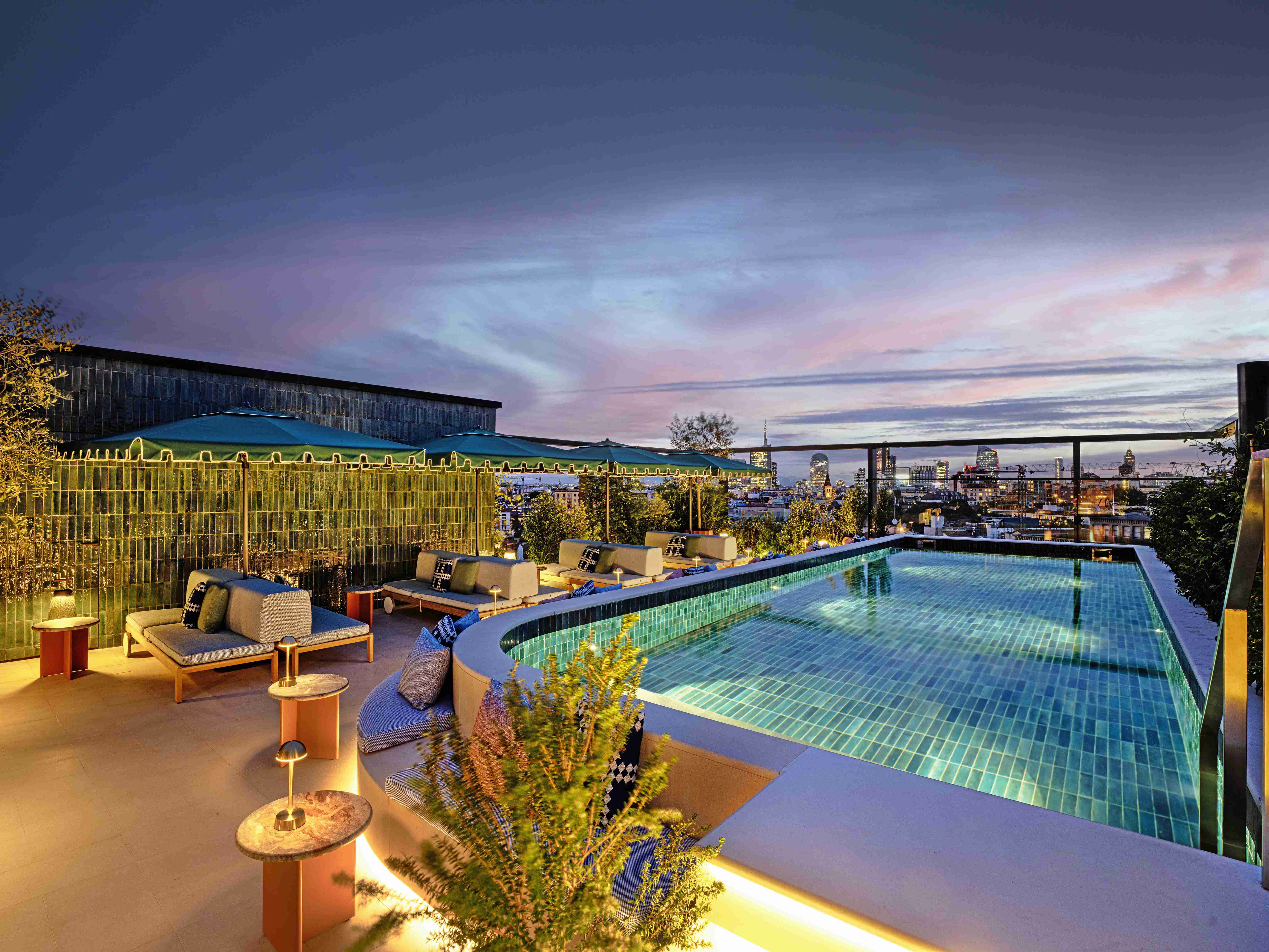 A hotel rooftop pool at night with soft lighting, lounge seating and city views