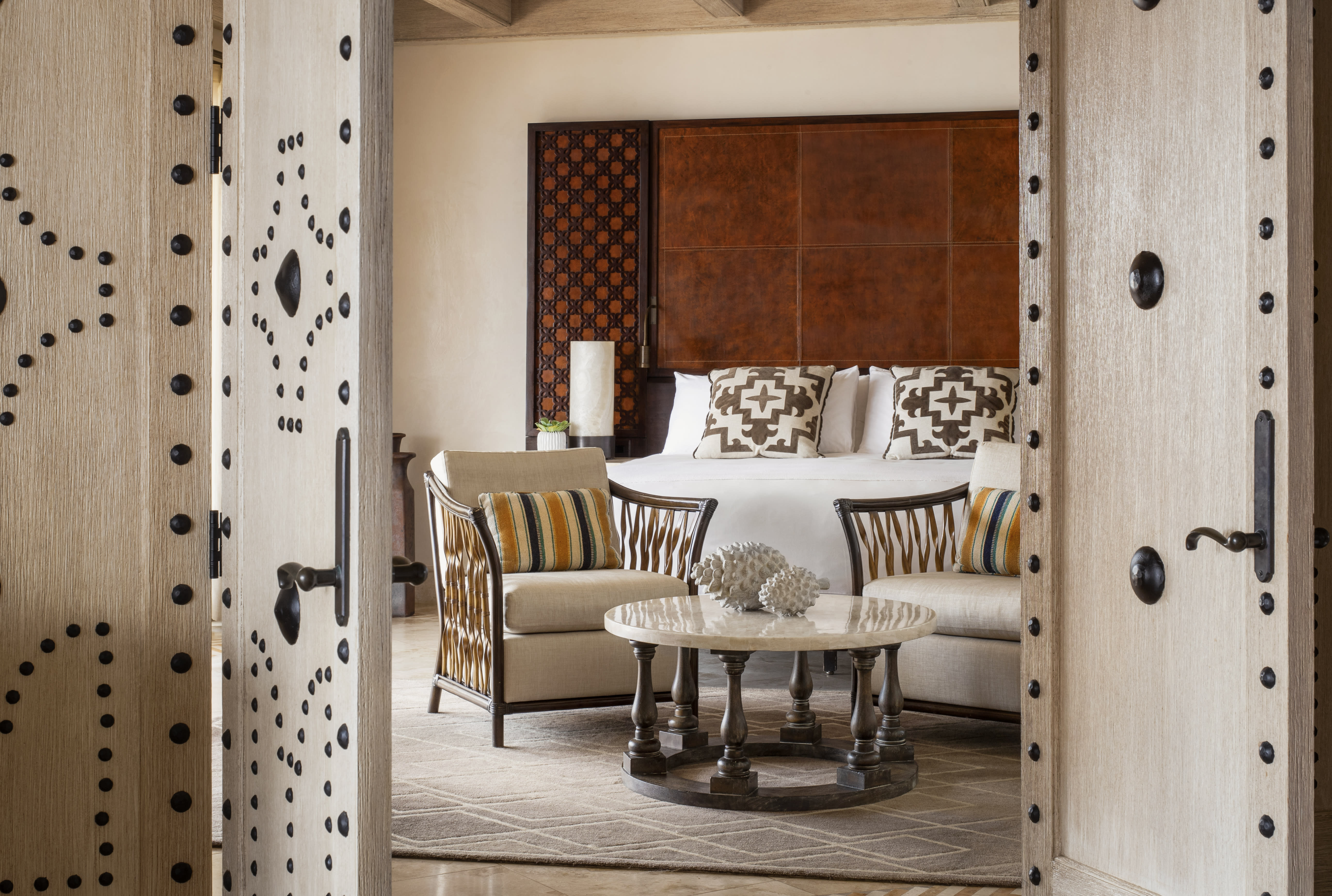 View through ornate wooden double doors into a luxurious hotel suite with a king bed, patterned pillows, upholstered armchairs, and a round coffee table topped with decorative coral.